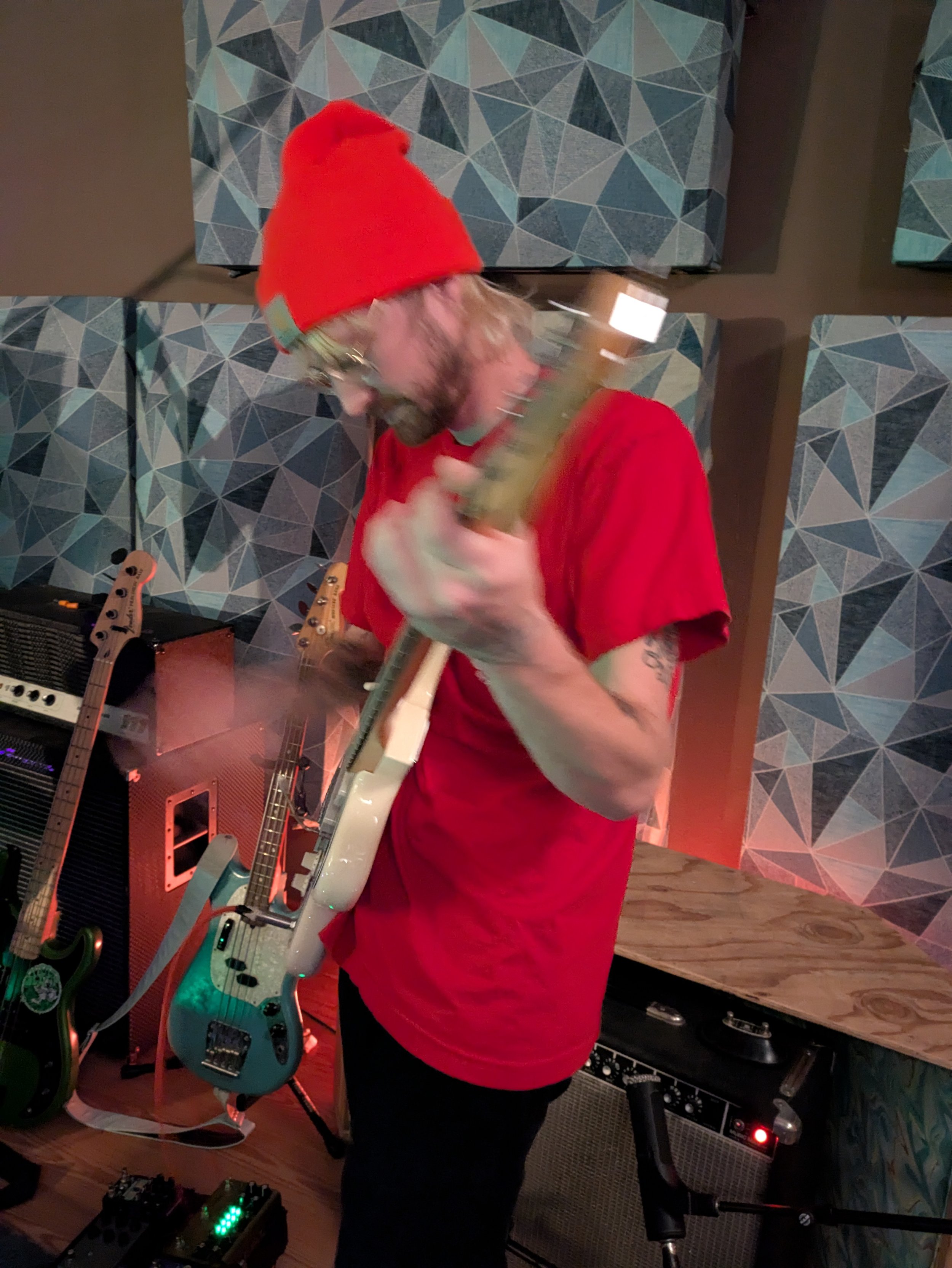 Person with long hair wearing a red beanie and a red t-shirt playing an electric guitar in a music studio, with various guitar pedals, amplifiers, and musical equipment around.