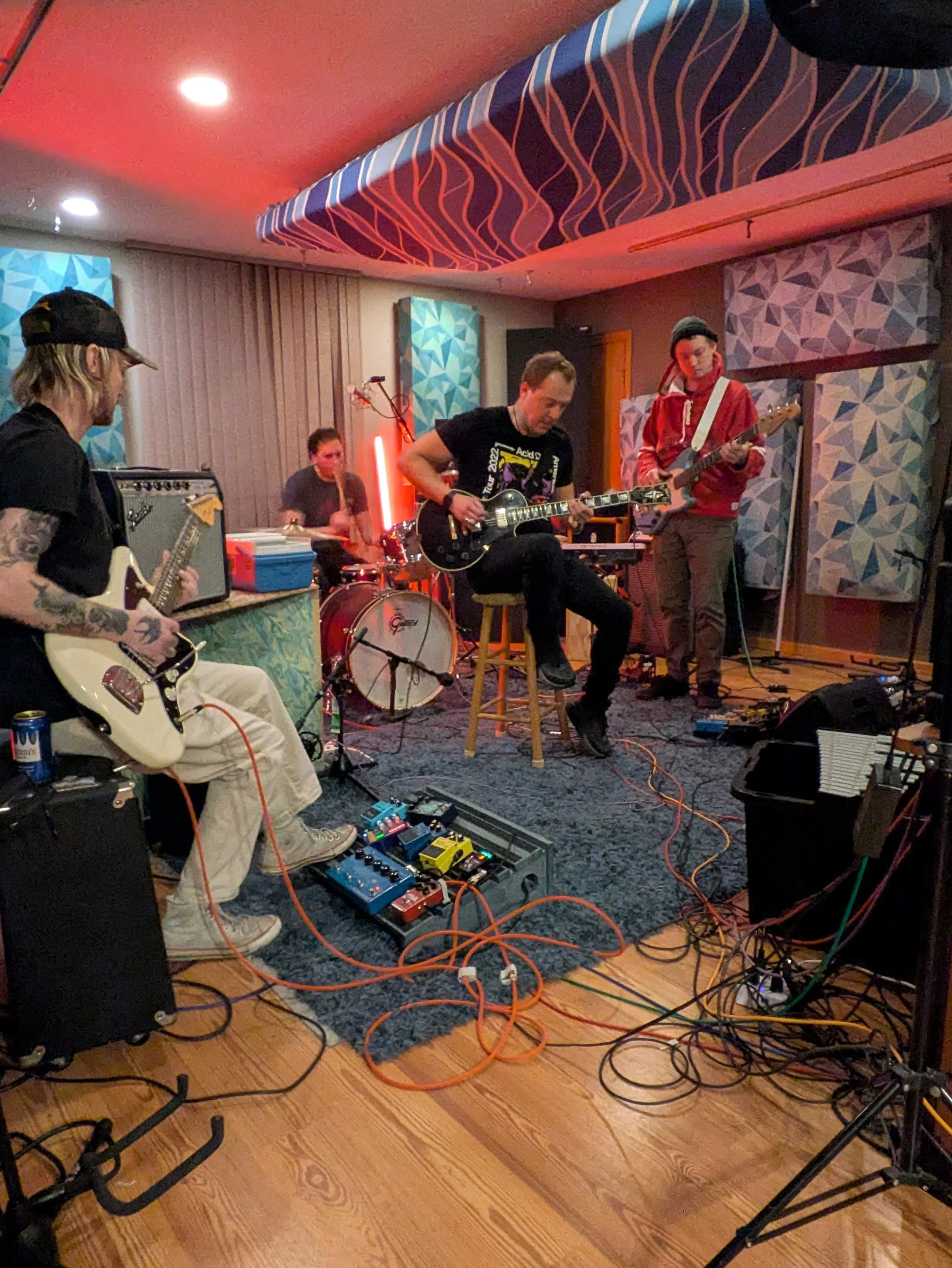 A band is performing in a room with acoustic panels on the walls and colorful carpeted ceiling. There are four musicians: a guitarist with tattoos wearing a black baseball cap, a drummer in the background, a keyboardist and guitarist seated, and anot