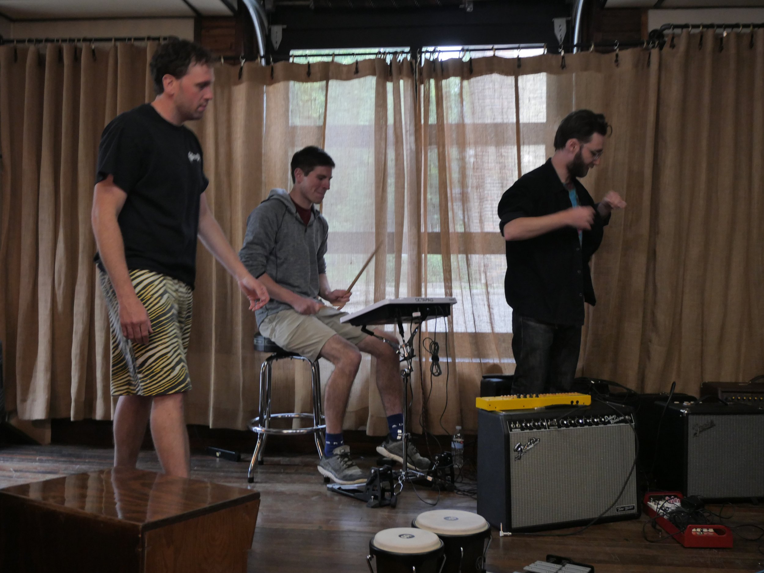 Three men are standing and sitting on stage in front of a beige curtain, with musical equipment like an amplifier, pedalboard, and bongos around them. The man on the left is wearing black and yellow striped shorts and a black shirt, the man in the ce