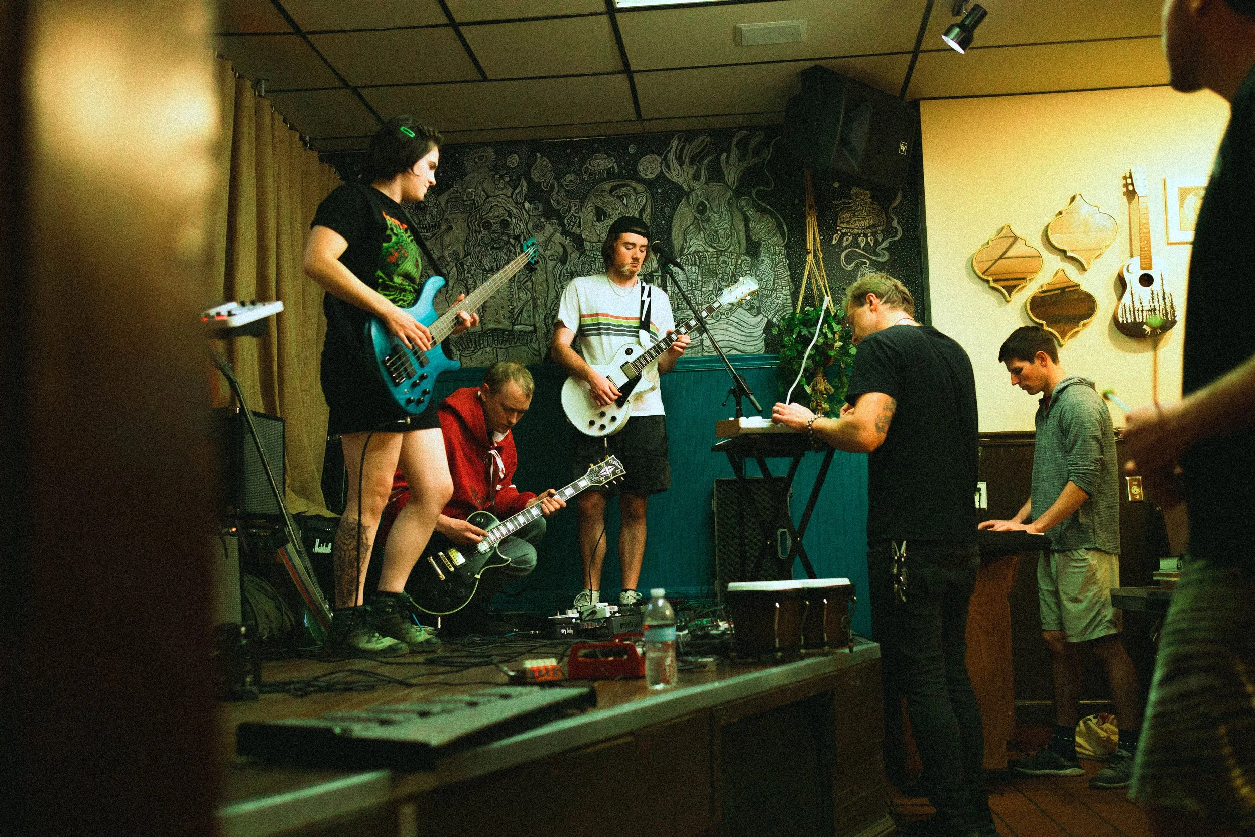 A band performing live on a small stage with several musicians playing guitars, keyboard, and singing, while an audience watches.