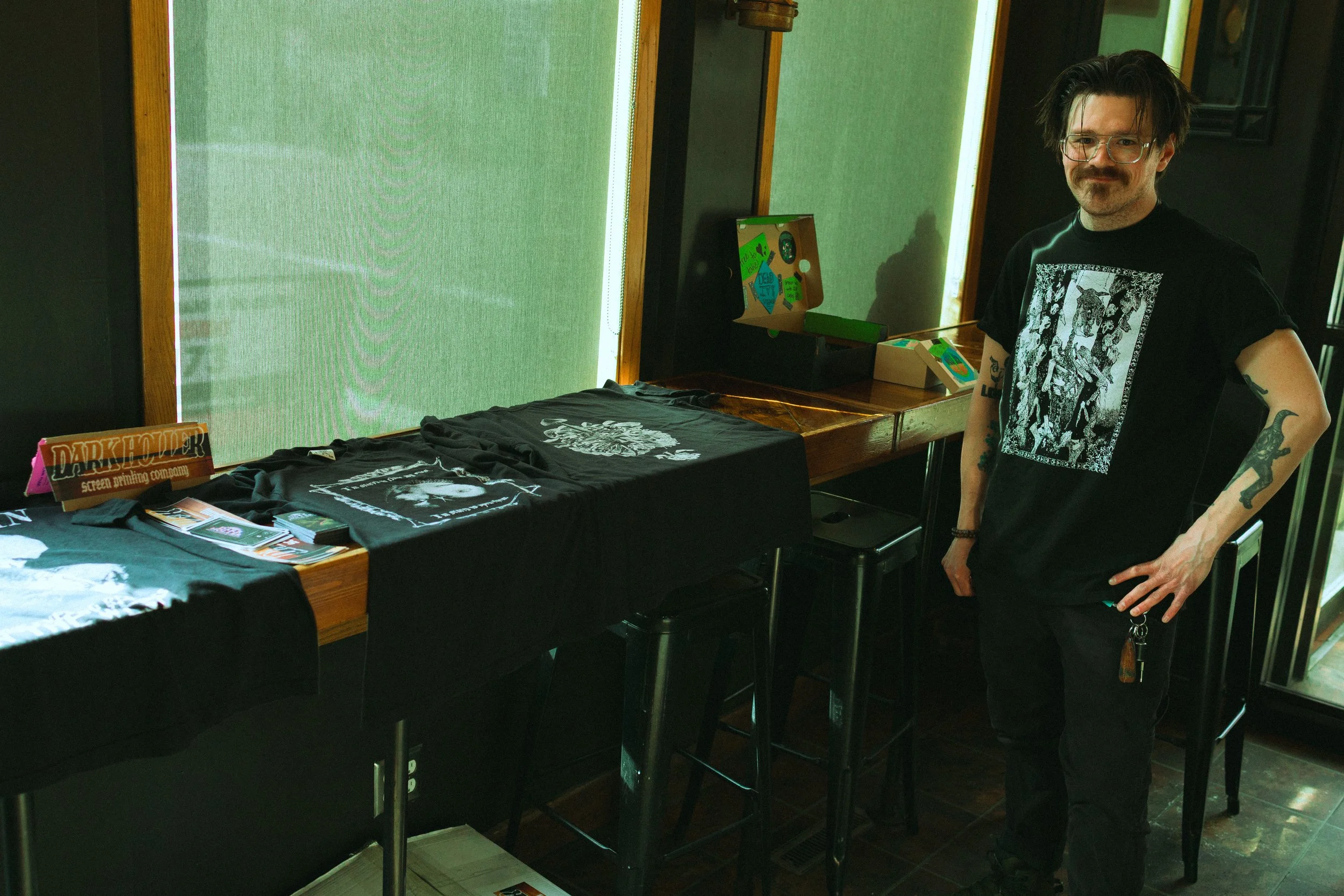 A man with glasses and tattoos wearing a black graphic t-shirt stands beside a table with black t-shirts featuring white designs, inside a room with green window blinds and framed pictures on the wall.