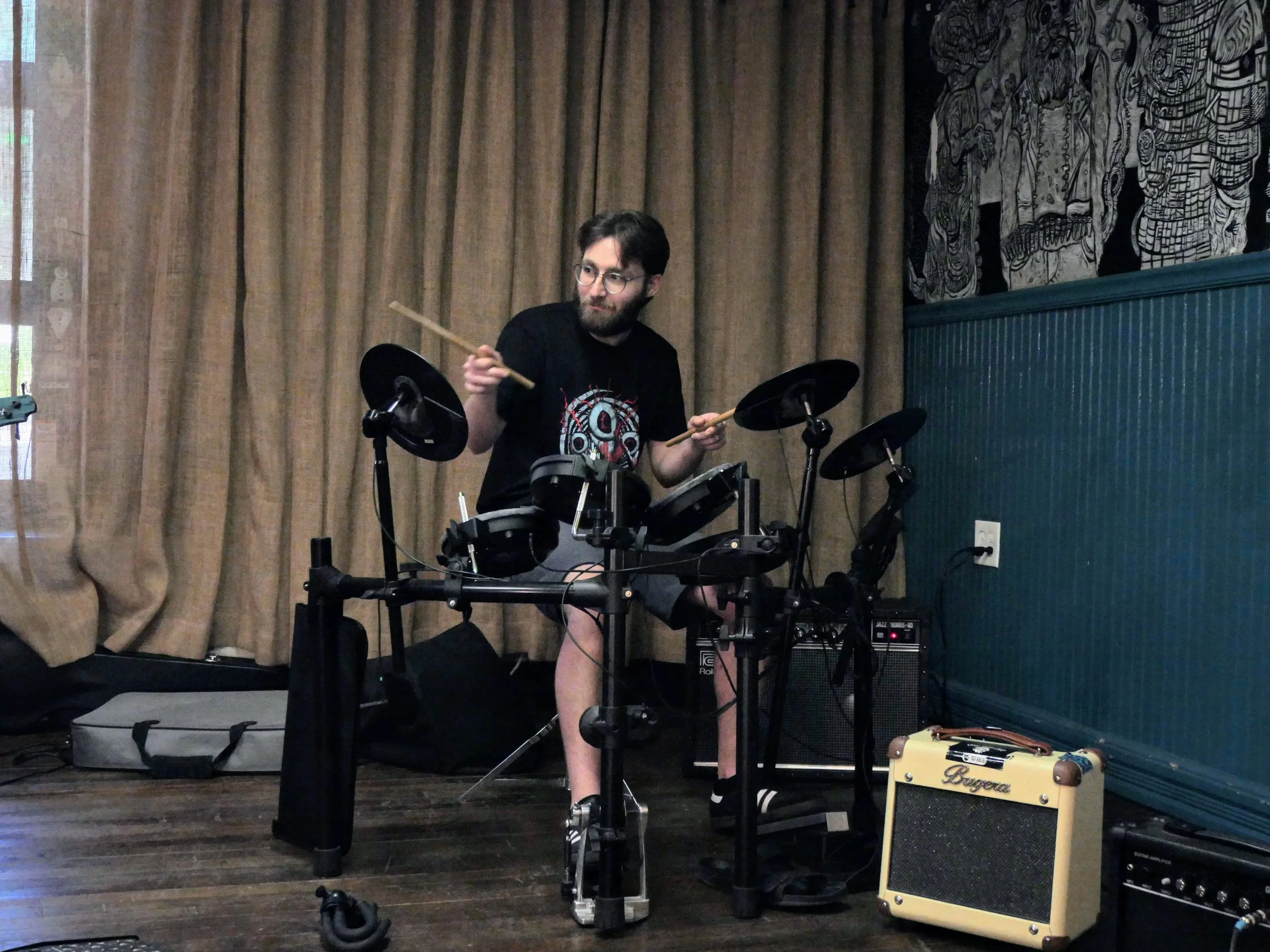 A man with glasses and a beard playing electronic drums indoors, with curtains in the background, a small amp, a guitar amplifier, and a yellow guitar amplifier nearby.