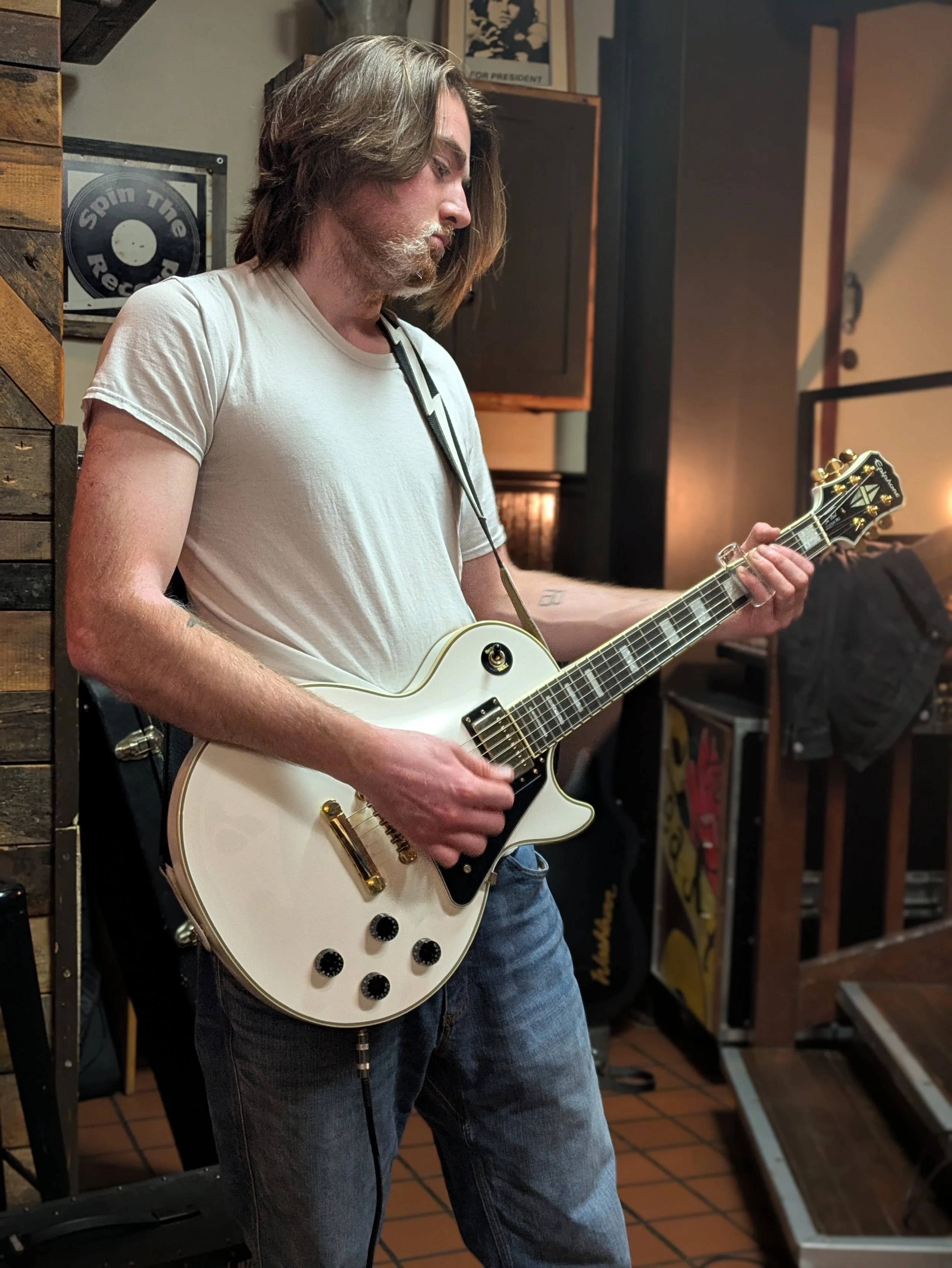 A man with shoulder-length brown hair and a beard playing a white electric guitar in a music studio, wearing a white t-shirt and jeans.