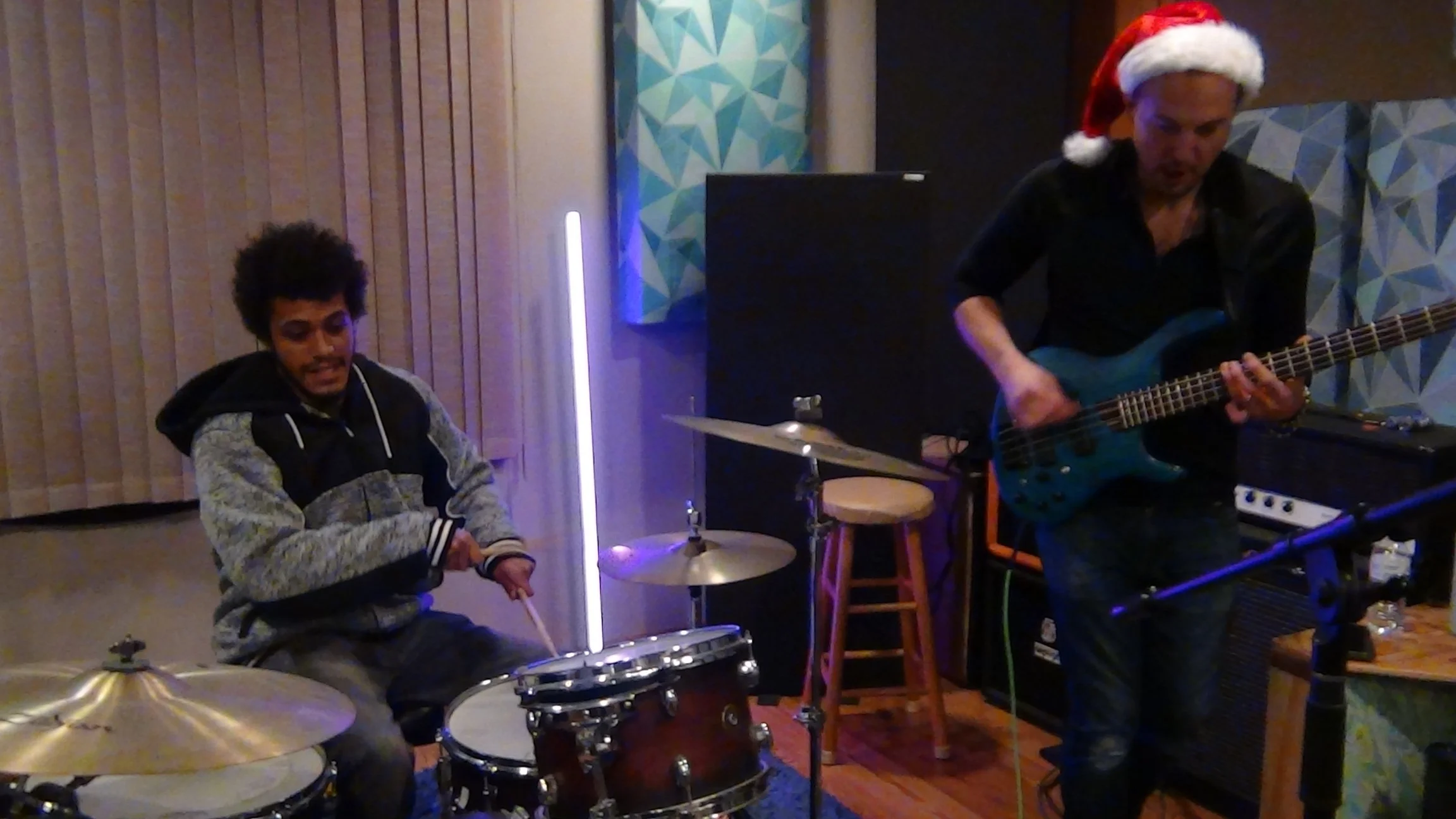 Two musicians playing instruments in a room decorated with geometric wall art; one is playing drums and the other is playing a bass guitar, the person with the bass is wearing a Santa hat.