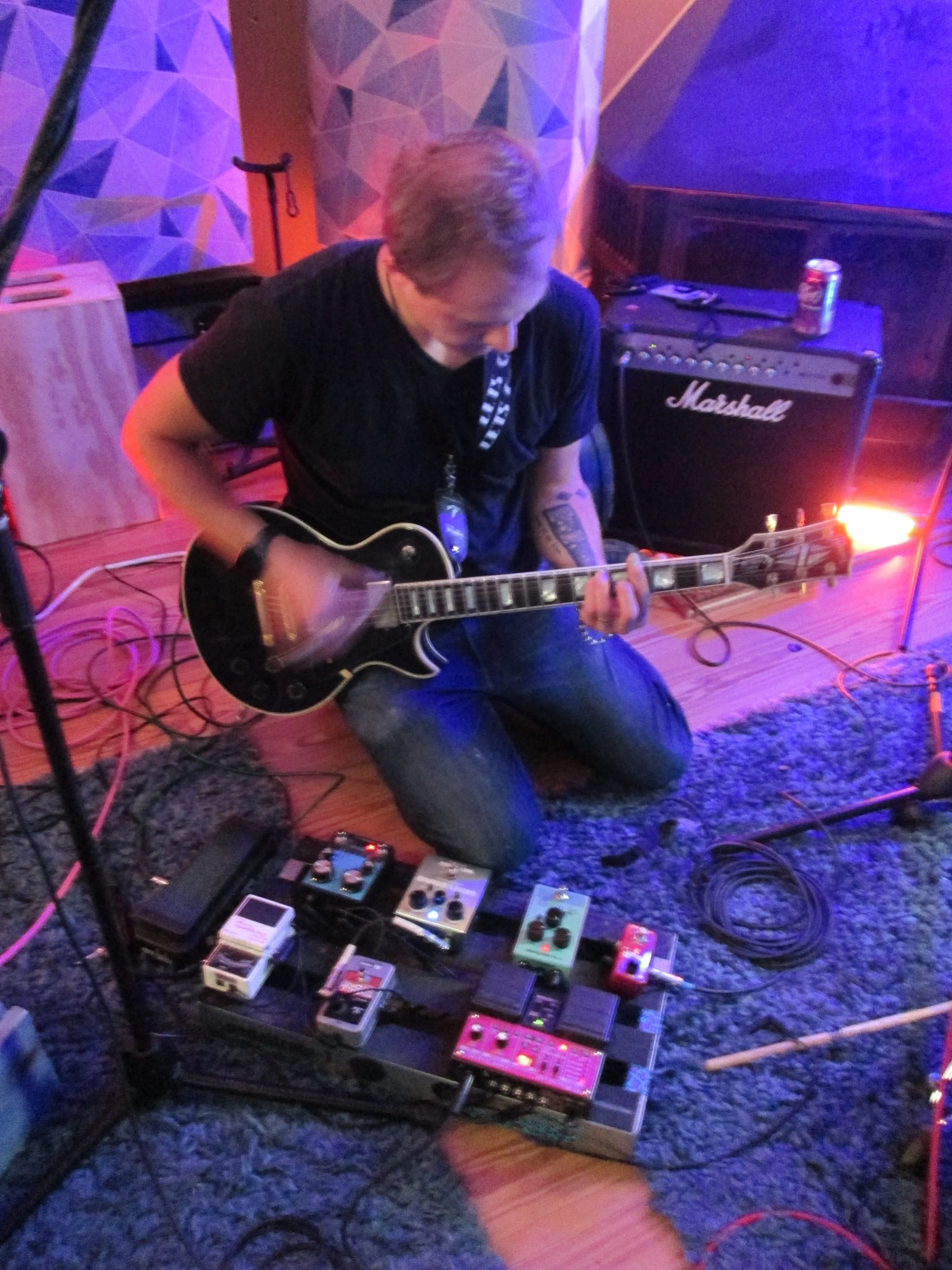 A person playing an electric guitar in a room with colorful lighting, surrounded by guitar effect pedals and an amplifier.