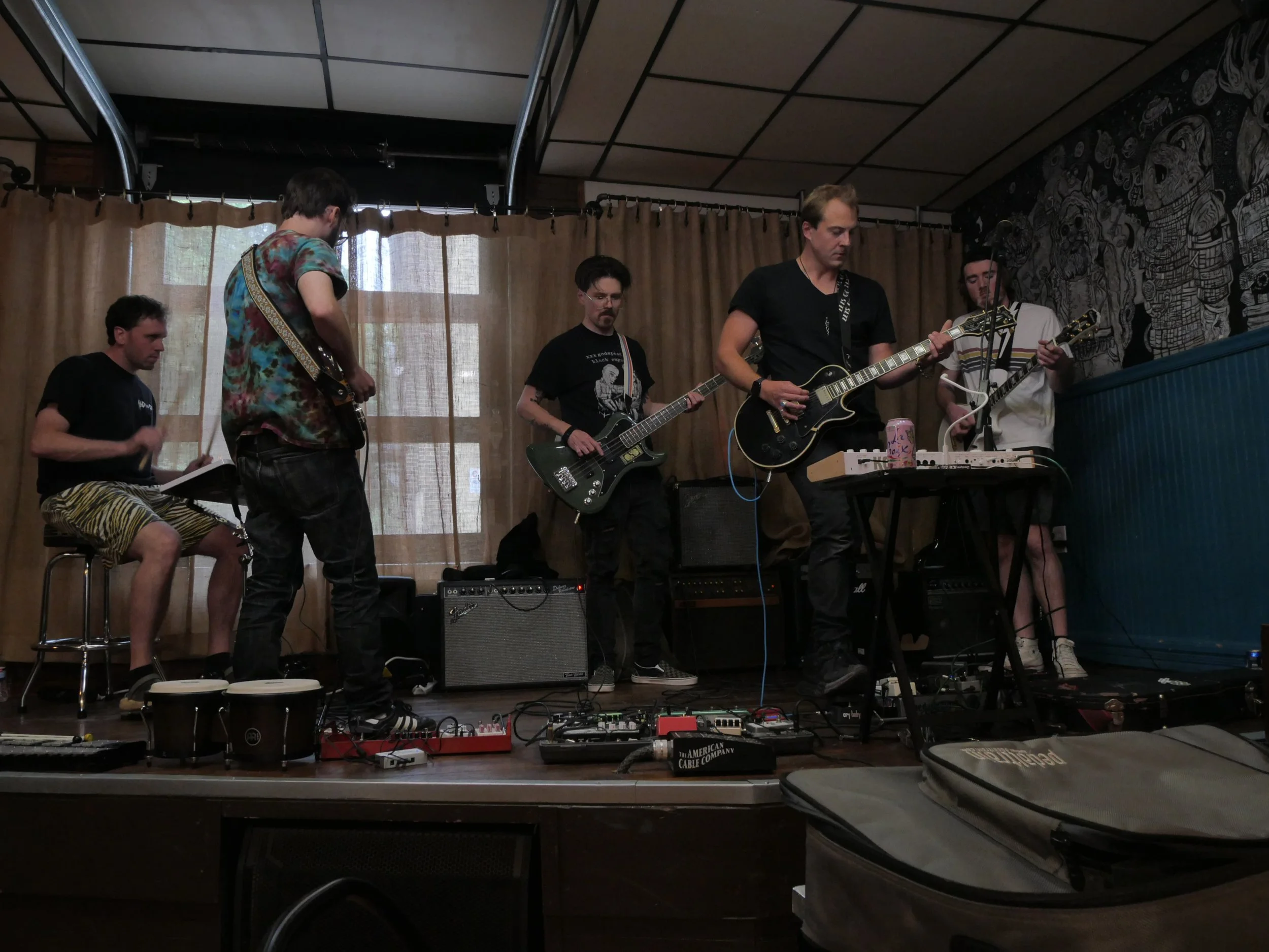 A band performing live on a small indoor stage with five members playing guitars and drums, surrounded by audio equipment and a patterned wall.