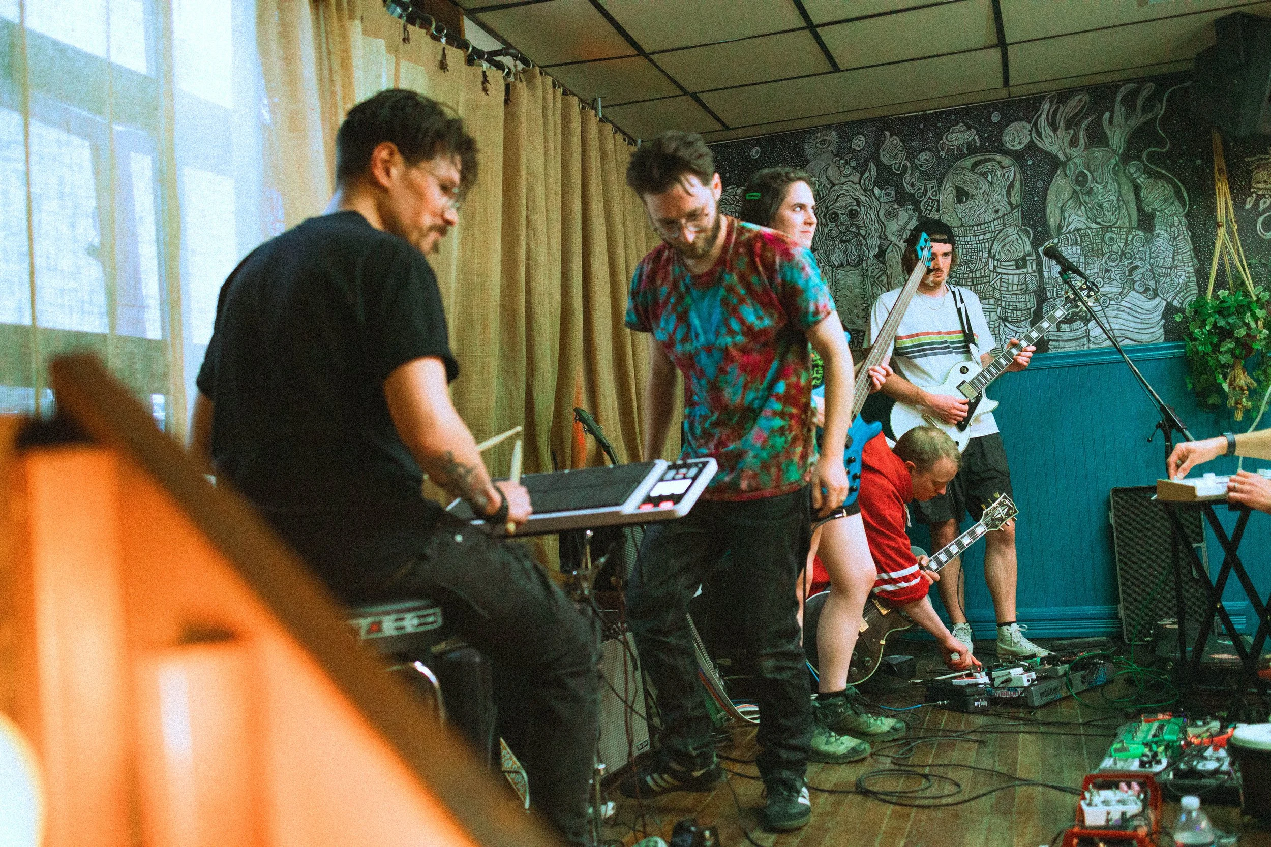 A group of young musicians and performers on a stage with musical instruments and pedals, preparing to perform or rehearse, with a detailed mural in the background.