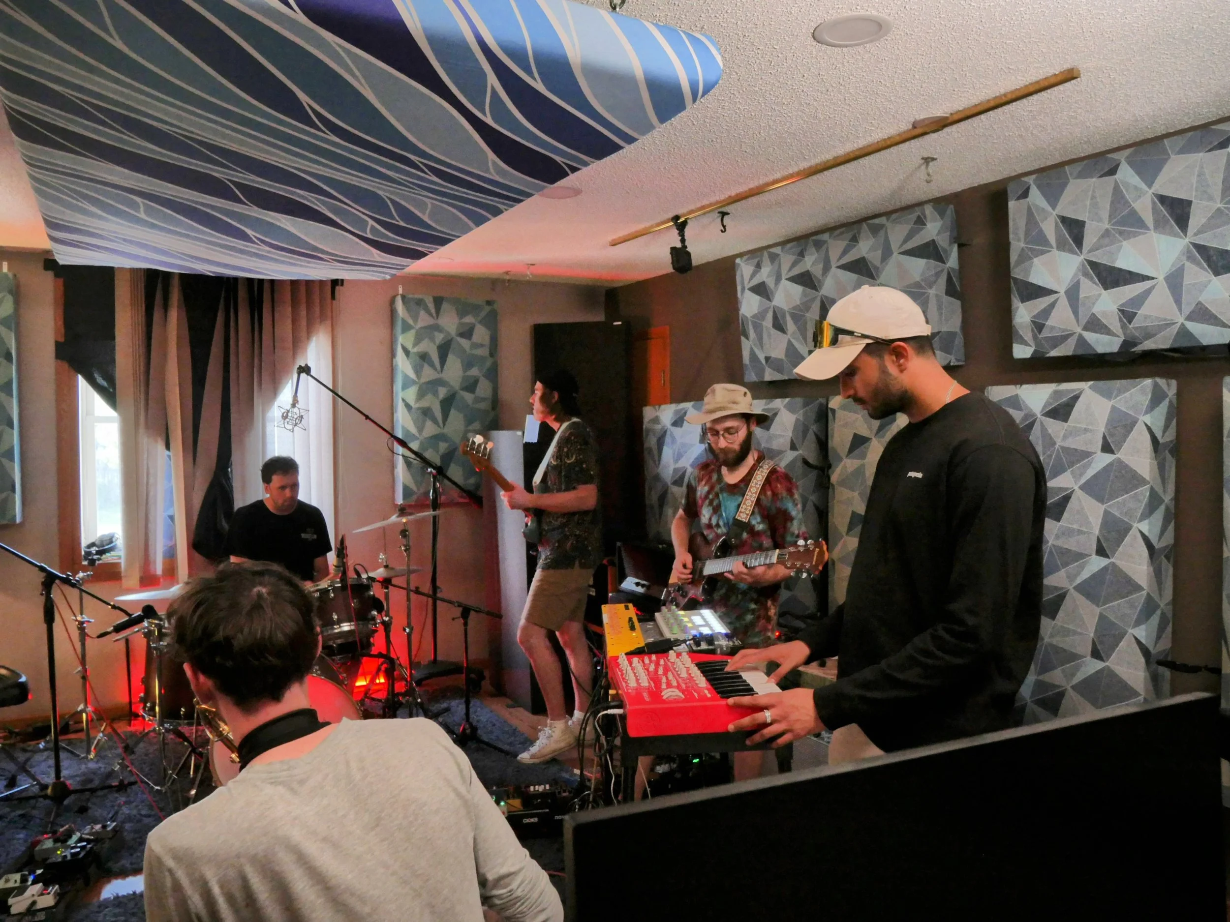 Musicians performing in a recording studio, including a guitarist, drummer, keyboardist, and others, with soundproofing panels on the walls and equipment around them.