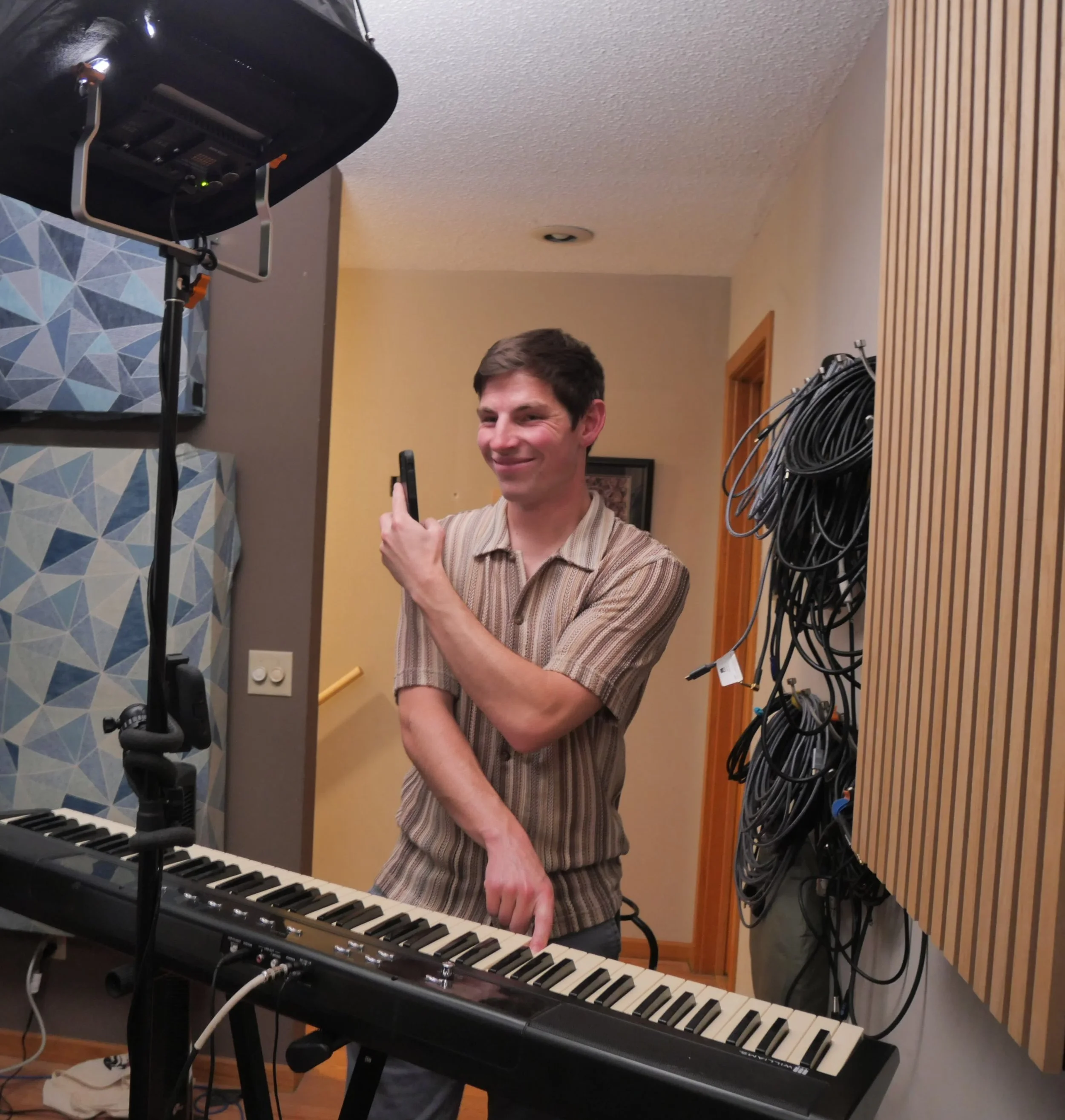 Smiling man with dark hair taking a selfie in a music rehearsal room, standing near a keyboard and music equipment.