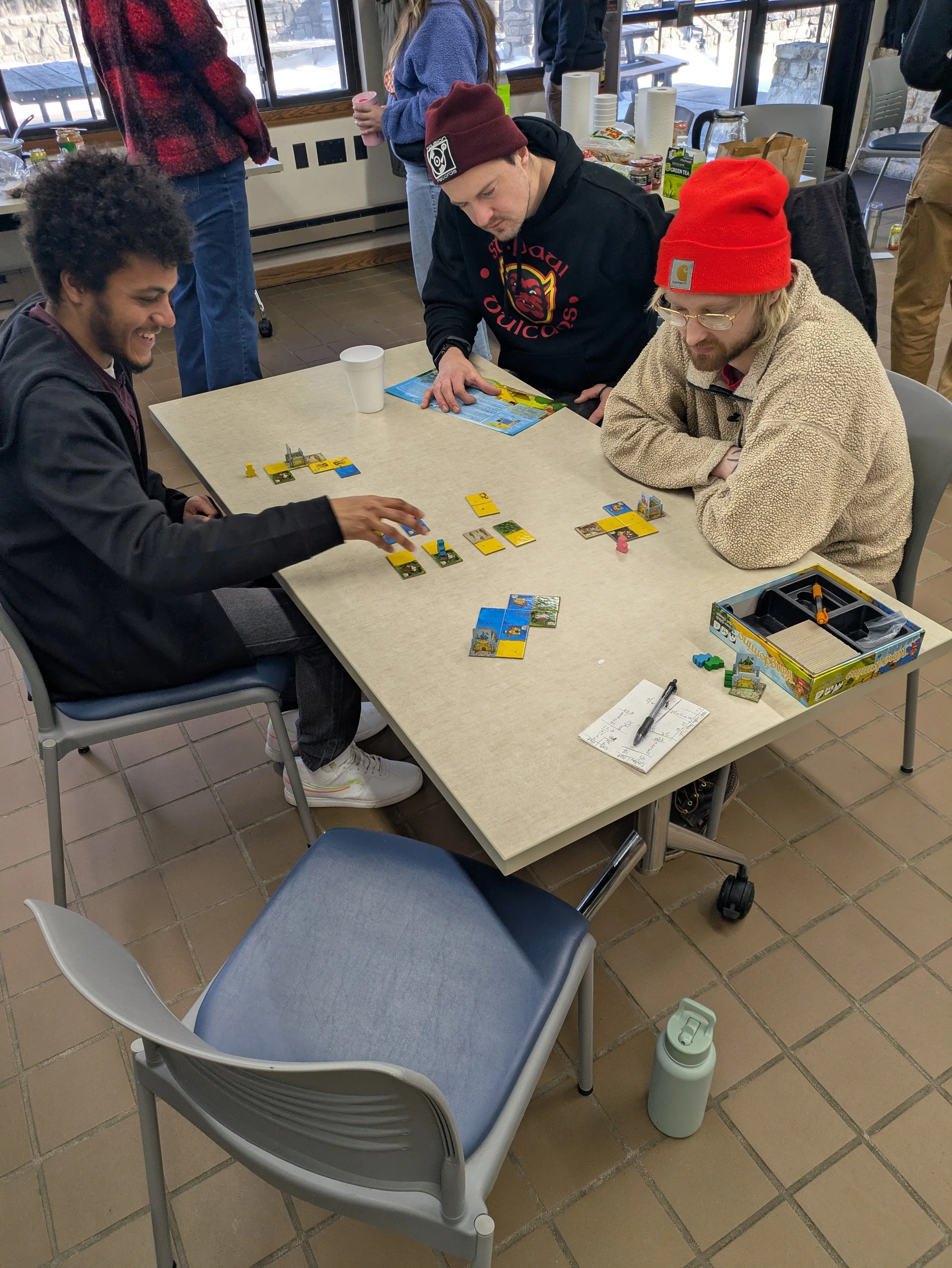 Three people are playing a game of Carcassonne at a table in a room with large windows. One person has curly hair and is opening a yellow and blue tile, while the other two are observing. The table has game pieces, cards, and a notebook nearby. A wat