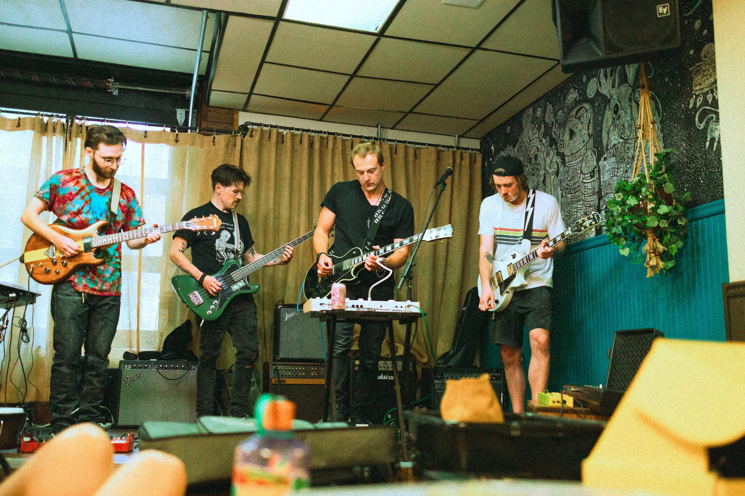 Four musicians playing guitars and keyboard on a small stage with a beige curtain background, a black and white wall mural, and a potted plant to the right. The scene appears to be at a live music performance or rehearsal in an indoor venue.