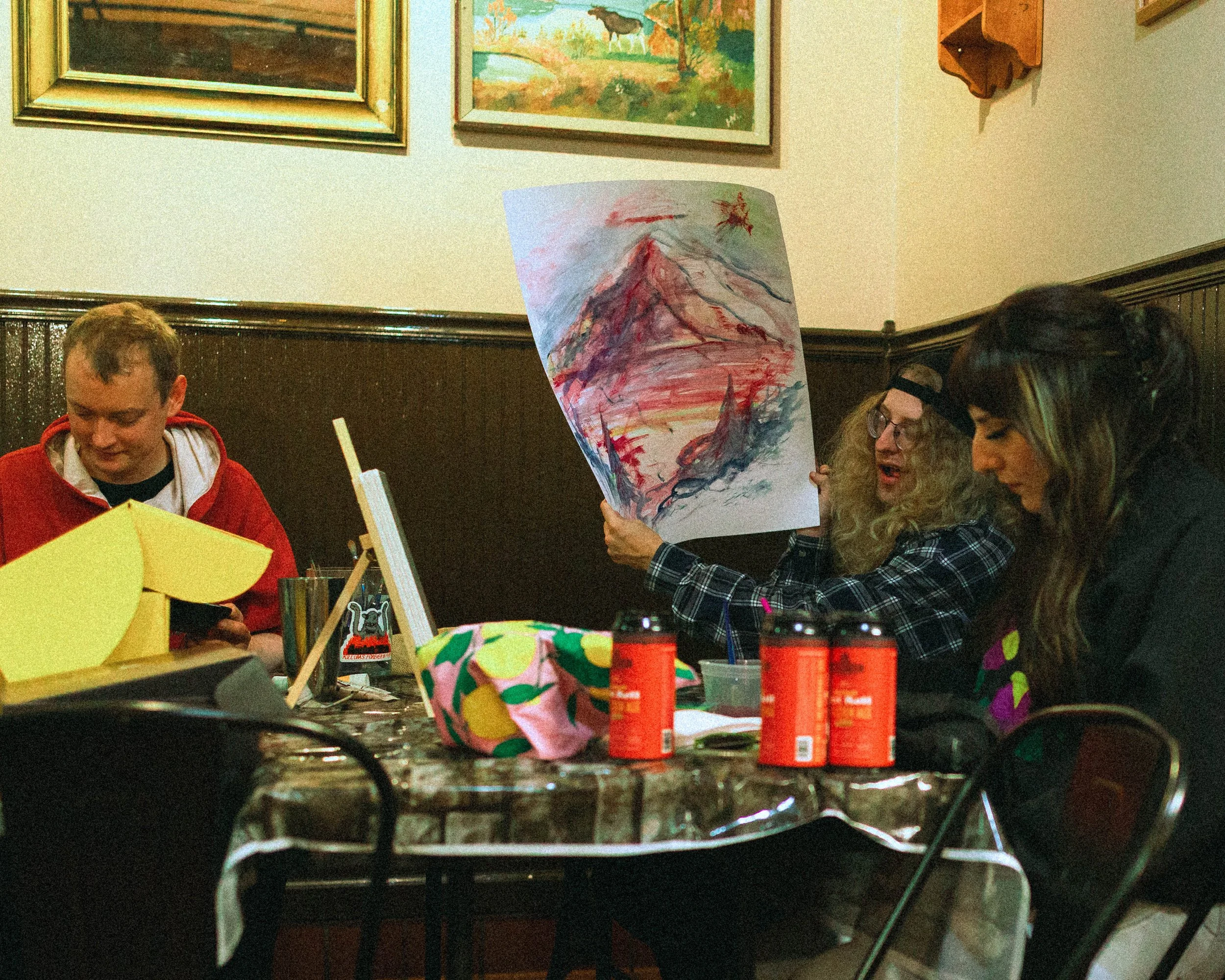 People sitting around a table in an art class, working on paintings; one person is showing a colorful mountain sketch on paper, and the scene features artwork and paintings on the walls.