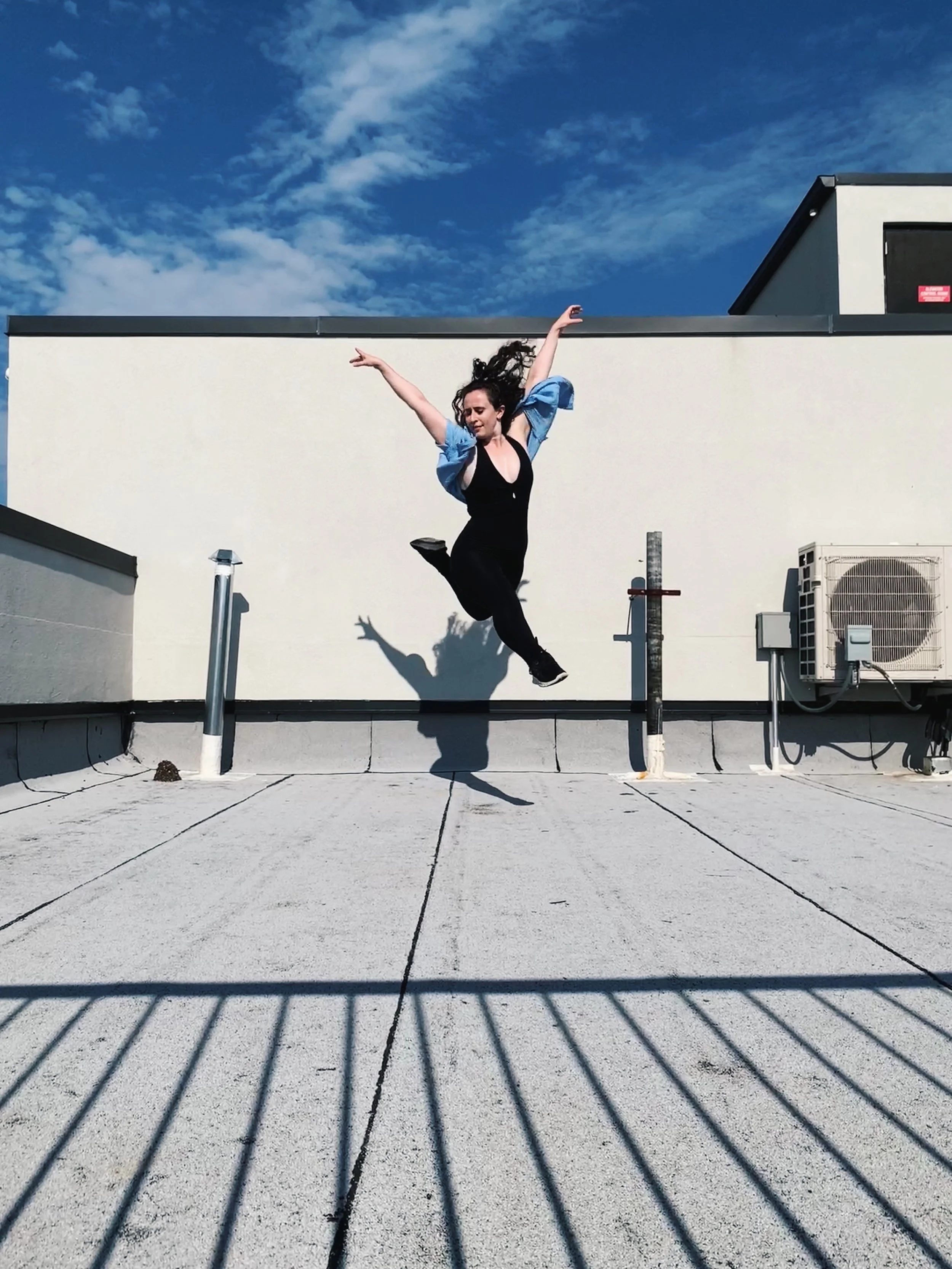A woman jumping on a rooftop with a white wall, blue sky, and clouds in the background. She is wearing black clothing and a blue jacket, with her arms raised in a dance pose.