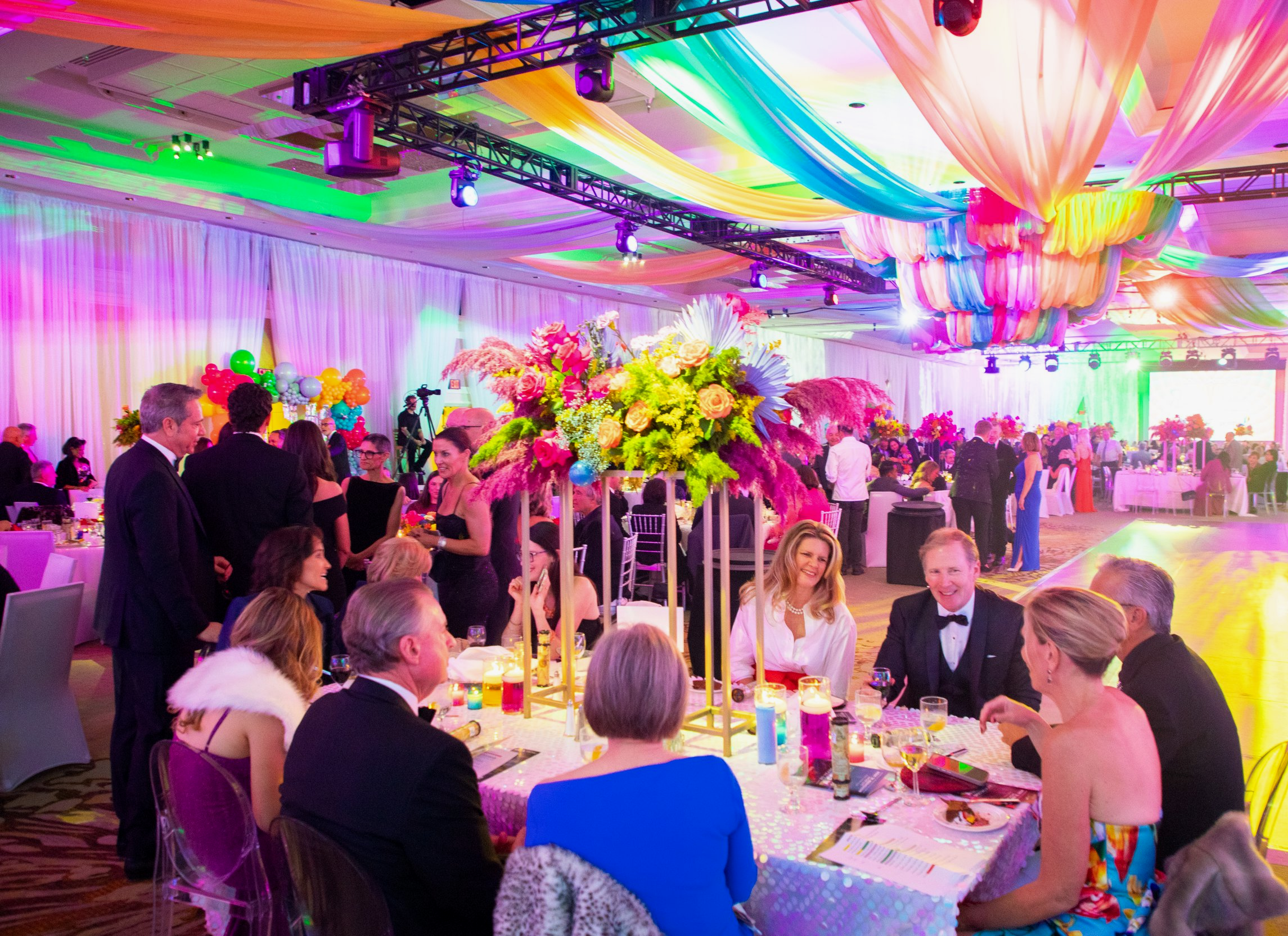 Guests dressed in formal attire socializing and dining at a colorful wedding reception with vibrant floral centerpieces, bright lighting, and draped ceiling fabric.