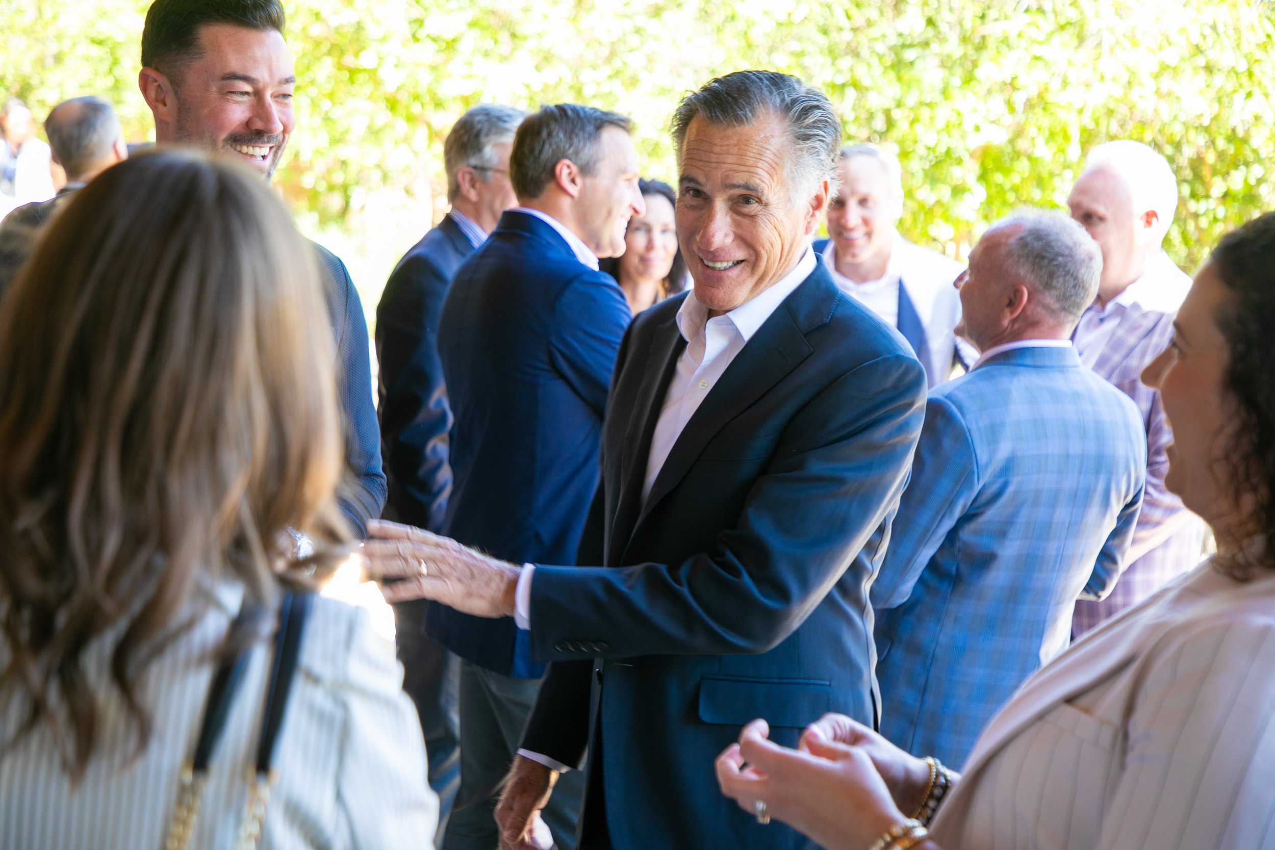 A group of people, some shaking hands, outdoors on a sunny day with trees in the background. The focus is on two men in suits smiling and greeting each other.