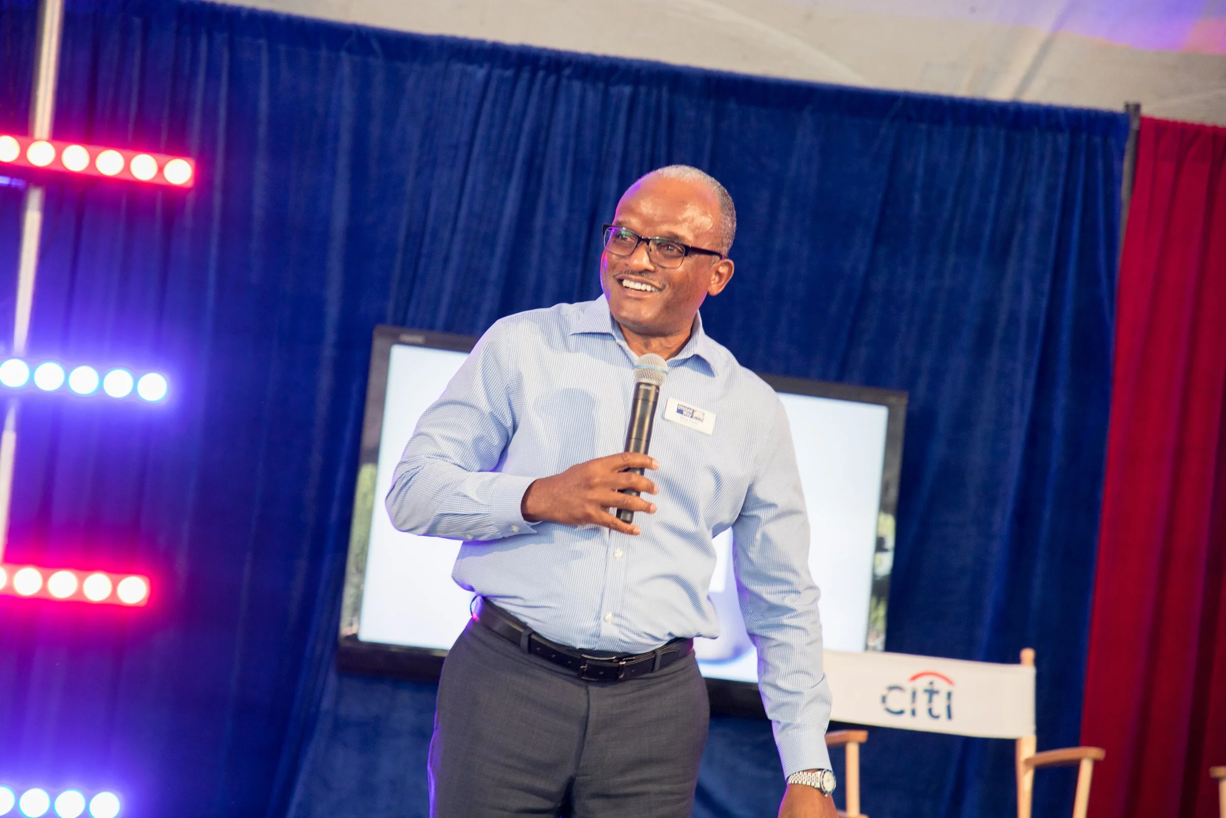 A man wearing glasses and a light blue dress shirt holding a microphone, standing on a stage with a blue curtain backdrop and red and blue lights, during a presentation or speech.