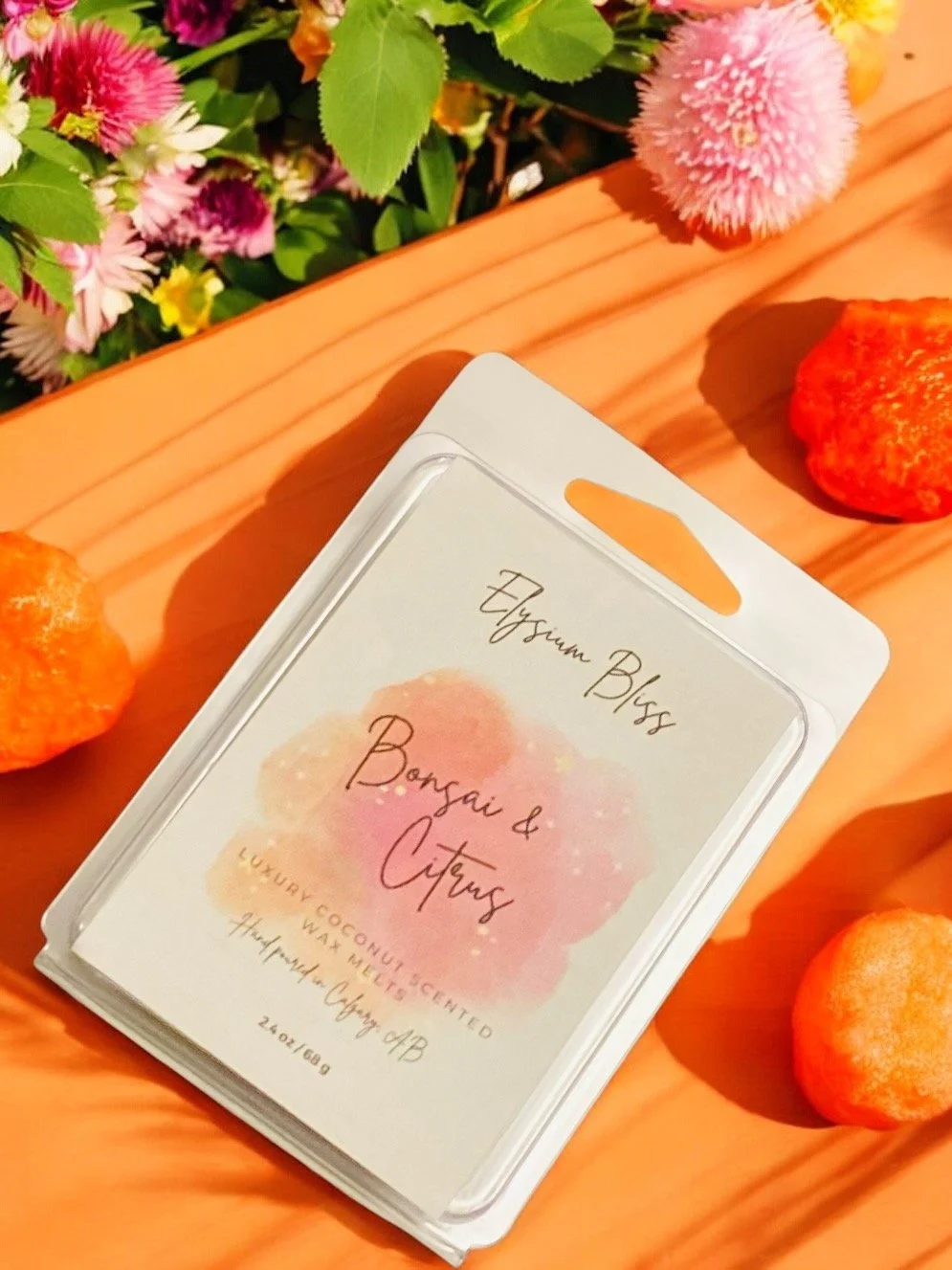 Packaging for a scented product with the label reading 'Elysian Bliss Bonsai & Citron, Luxury Coconut Scented Wax Melts.' The packaging is placed on a wooden surface surrounded by pink, purple, and yellow flowers and three small oranges or tangerines.