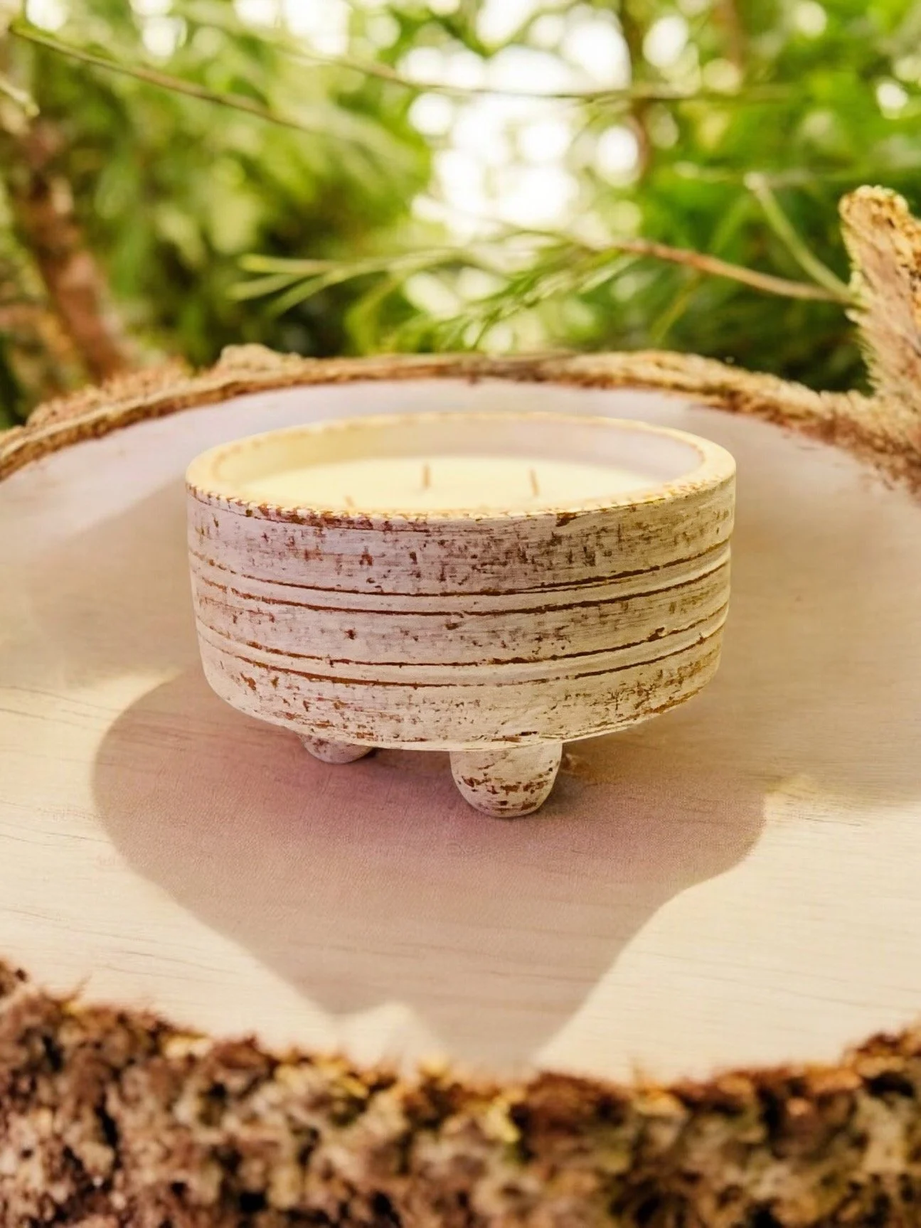 White rustic candle holder with three legs, placed on a wooden surface with a blurred natural outdoor background.