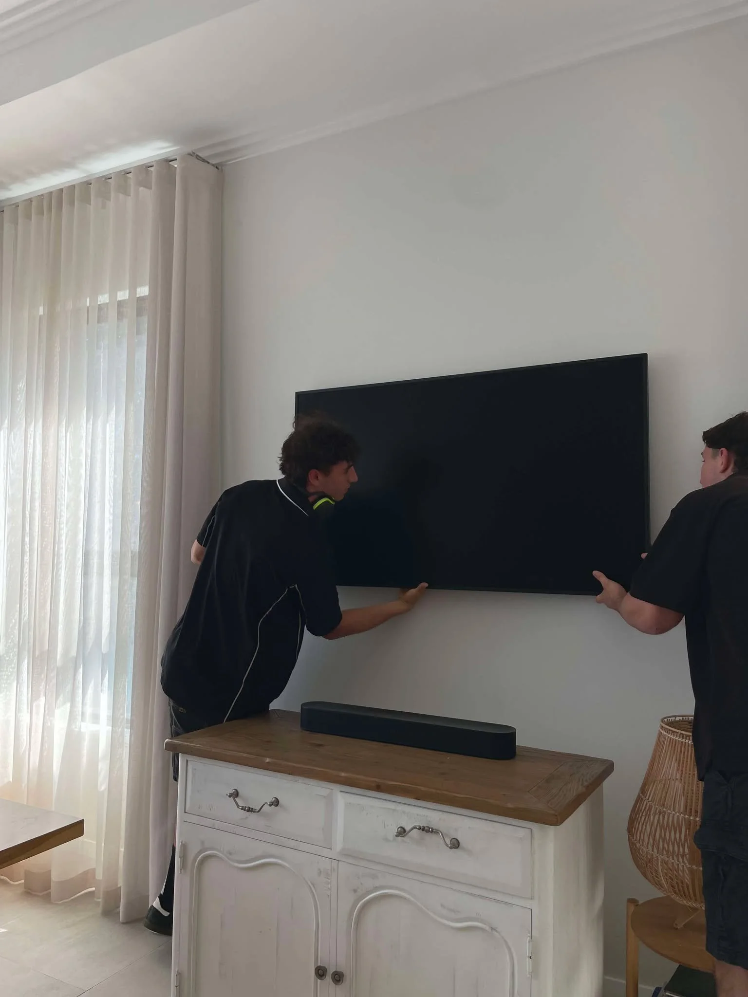 Two young men are mounting a flat-screen TV on a white wall in a living room, with a soundbar placed on a wooden cabinet below the TV.