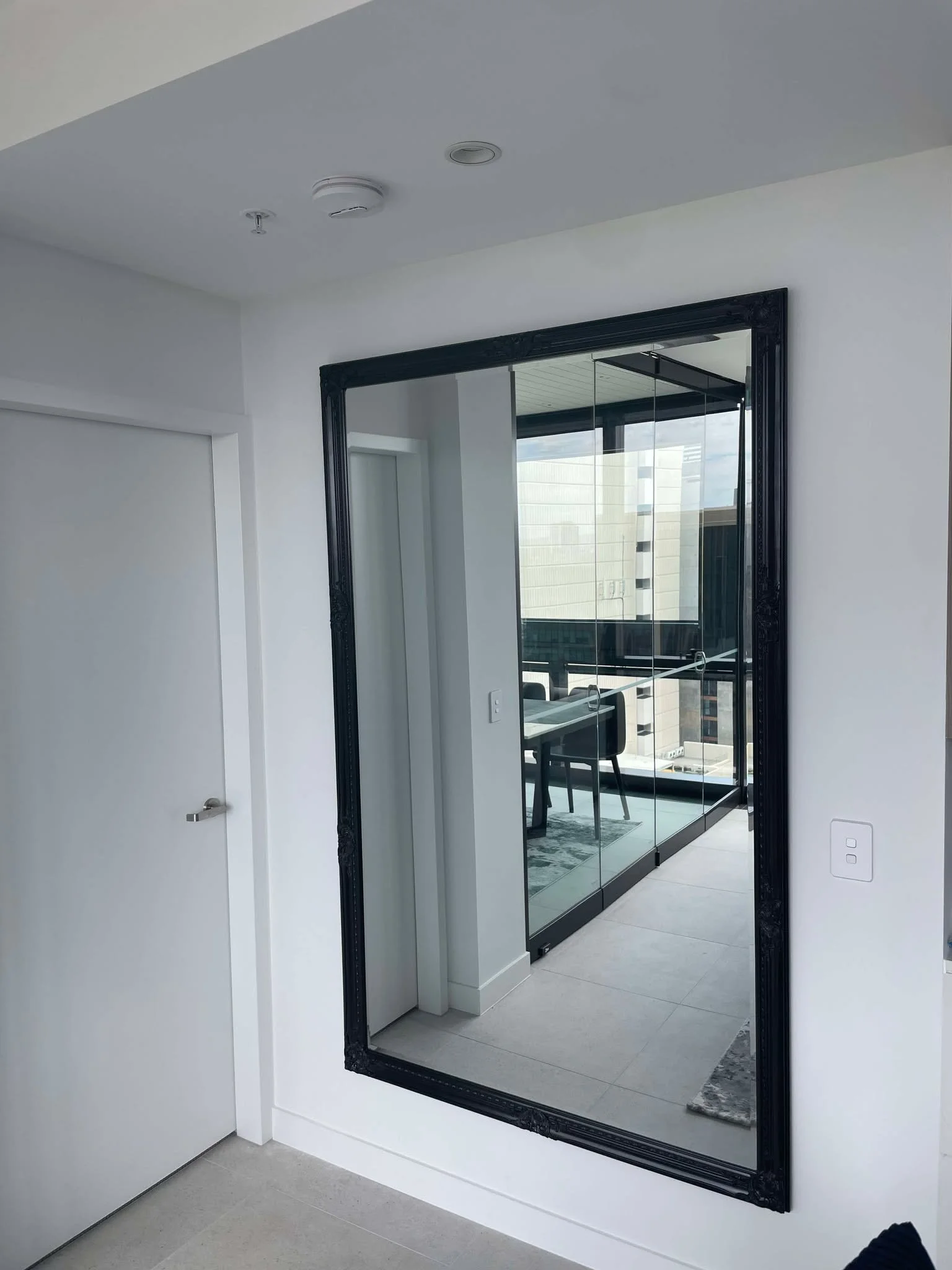 A large framed mirror on a white wall in a modern room reflecting a balcony with view of nearby buildings and furniture.