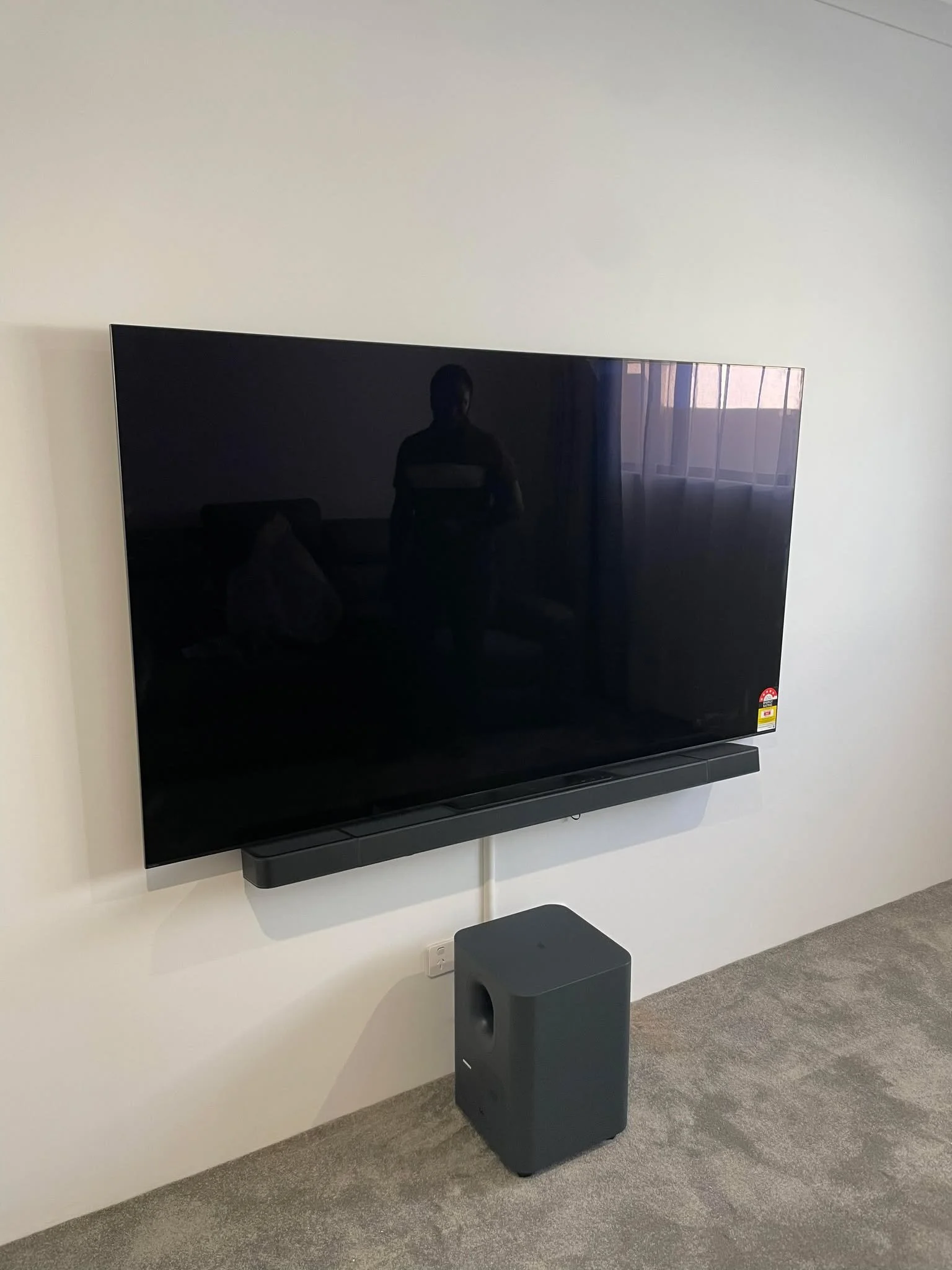 Flat-screen television mounted on a white wall with a black soundbar underneath. A gray subwoofer speaker is on the floor below the TV. The reflection of a person taking the photo and a window with curtains can be seen on the TV screen.