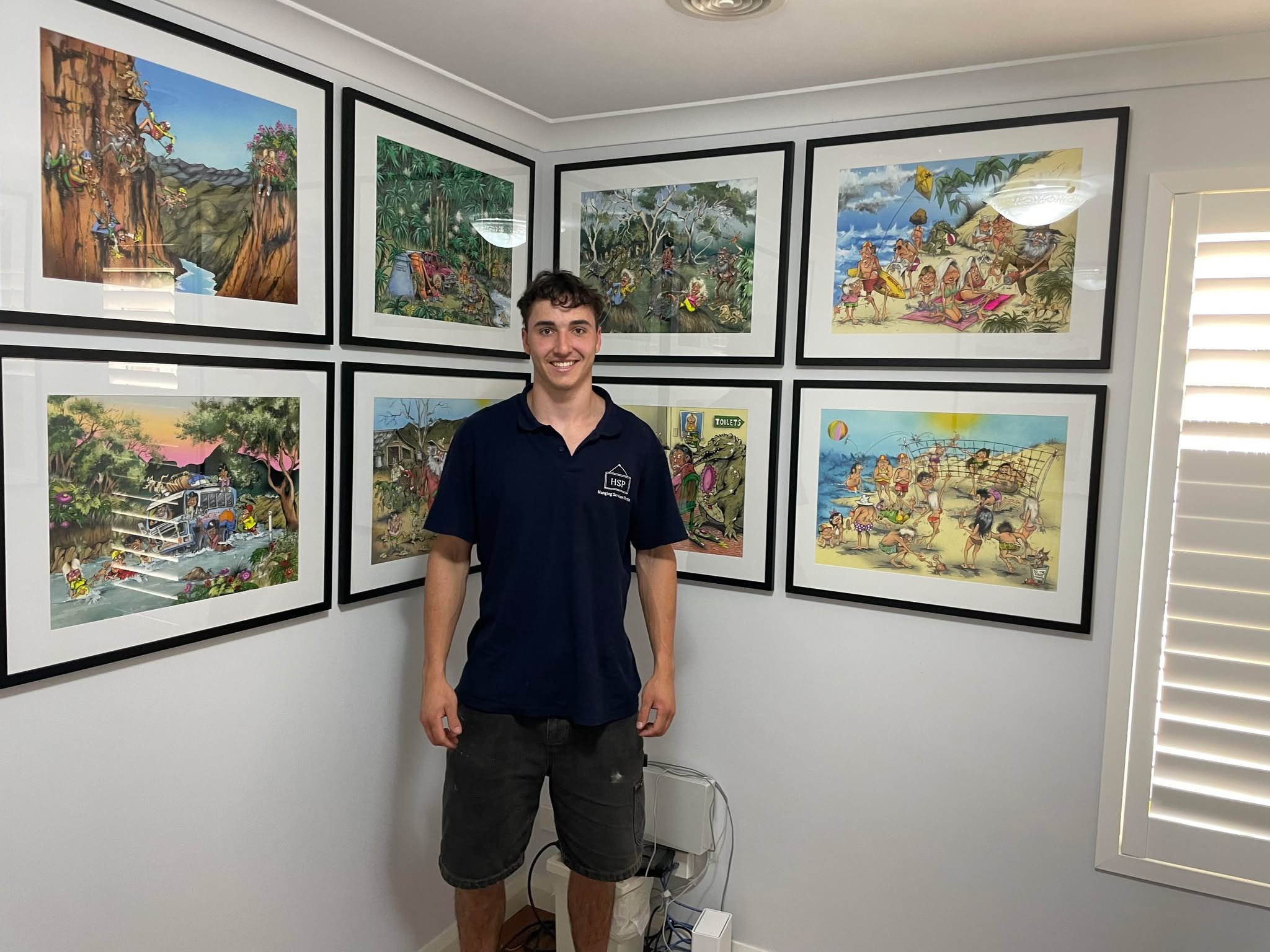 A young man smiling standing in front of a wall displaying framed colorful art prints, with a window with white shutters on the right side.
