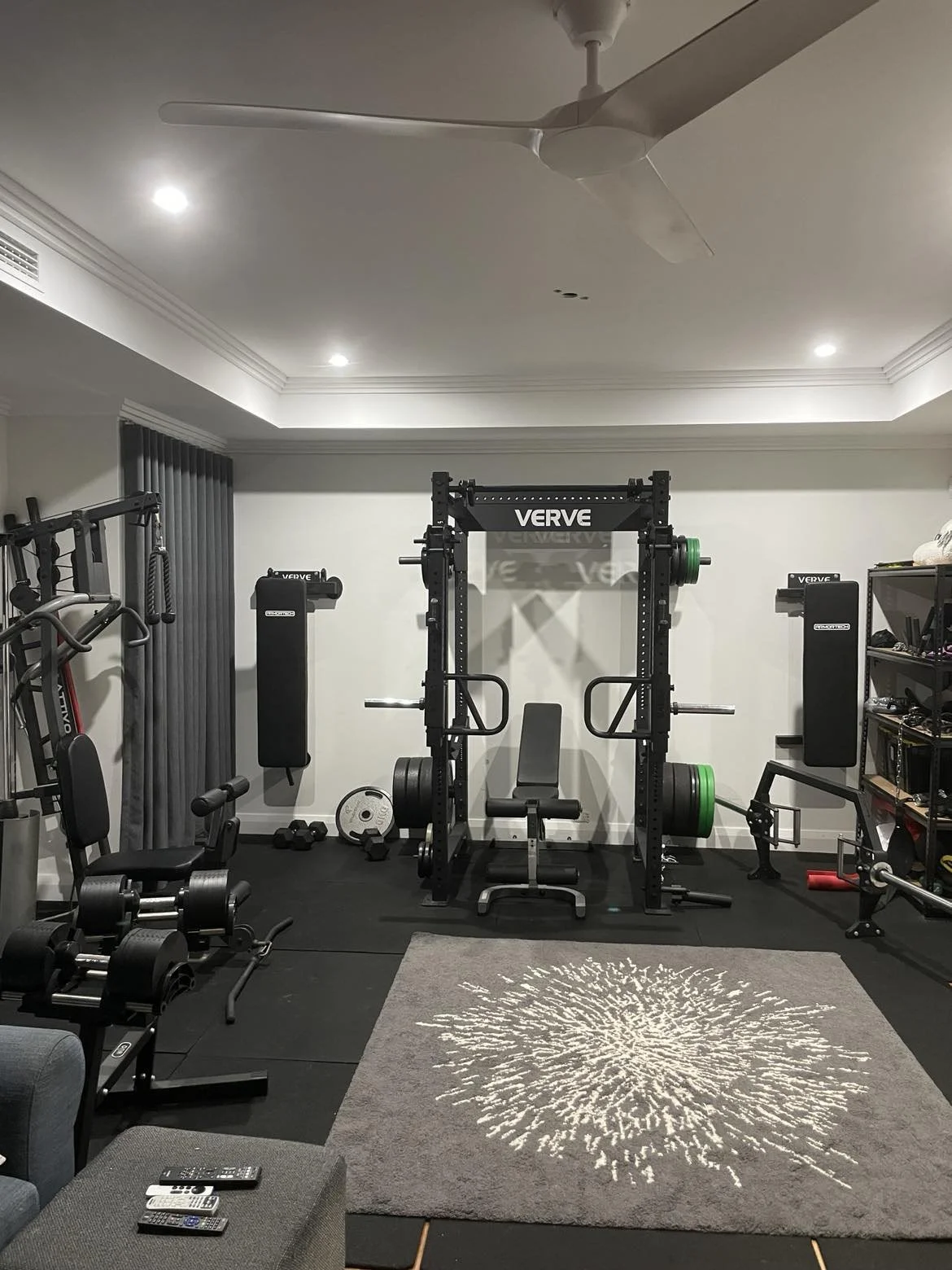 Home gym with a power rack, weight plates, a bench, dumbbells, and various exercise equipment. Centered on the wall is a black power rack labeled "VERVE" with a bench inside it. To the left are weight plates and a barbell on the floor, and to the right are shelves with more workout gear.
