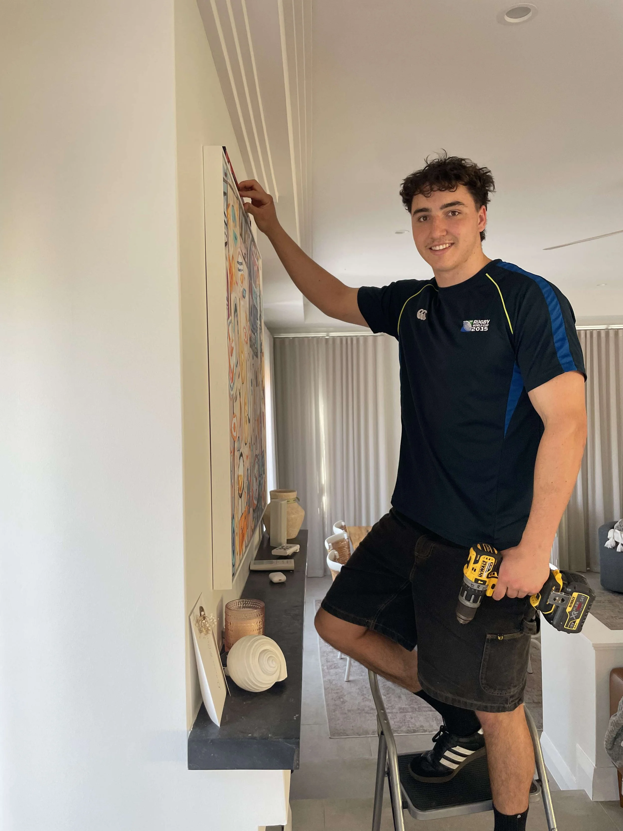 A young man standing on a step ladder in a living room, holding a drill and reaching up to a colorful abstract painting on the wall.