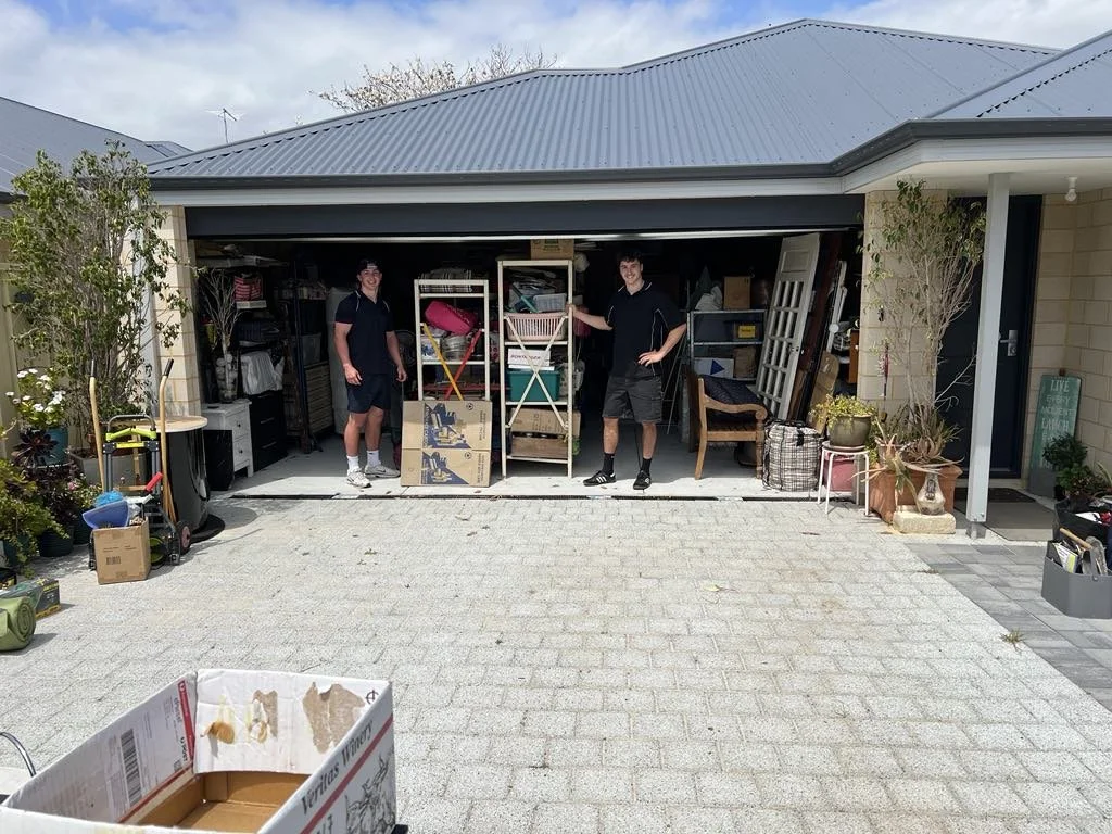 Two young men standing inside an open garage filled with various household items and furniture, with a paved driveway in front and potted plants on the sides.