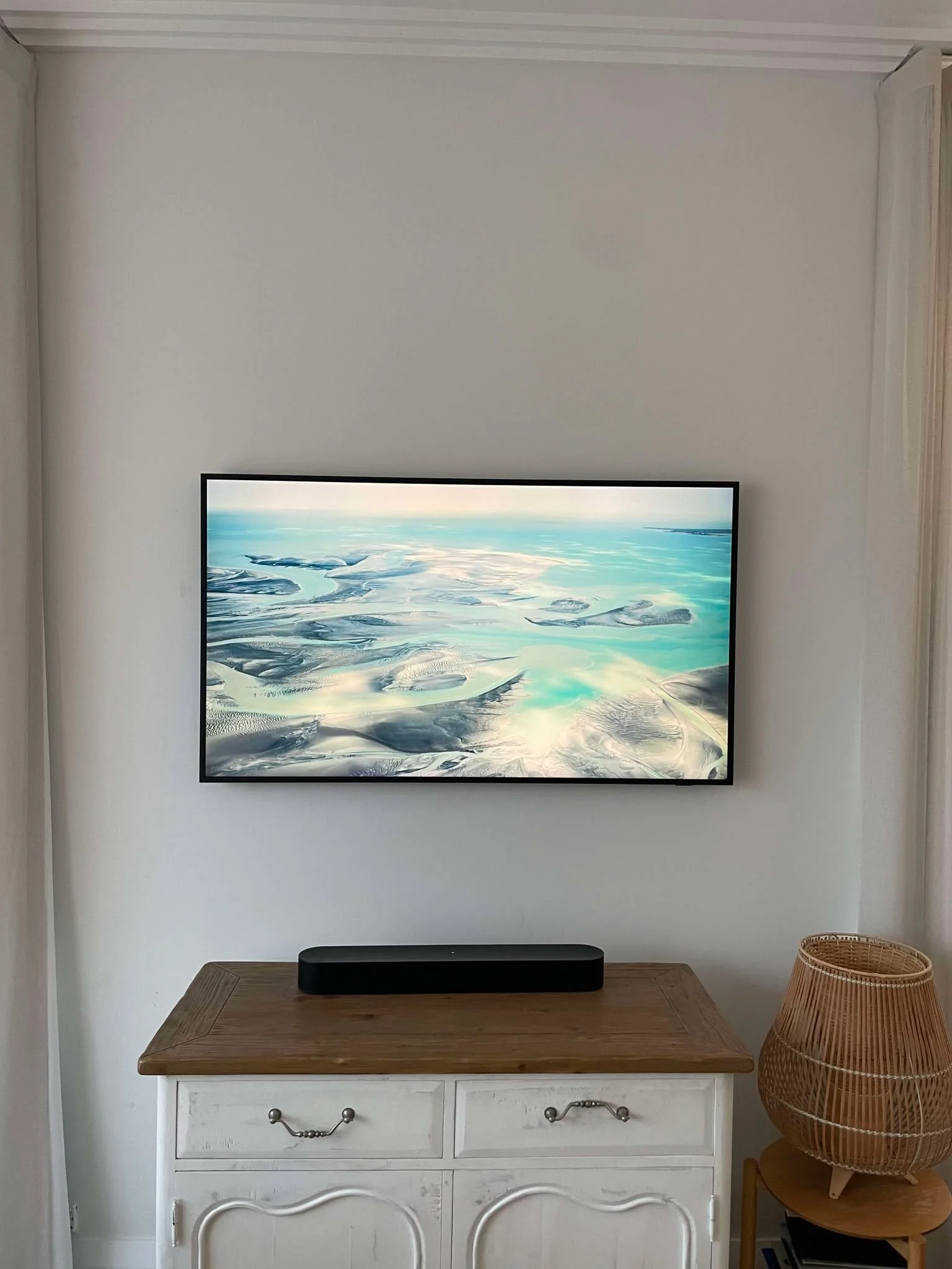 Living room with wall-mounted flat-screen TV displaying a landscape image, a white wooden console table with a black soundbar on top, and a wicker lampshade on a side table to the right.