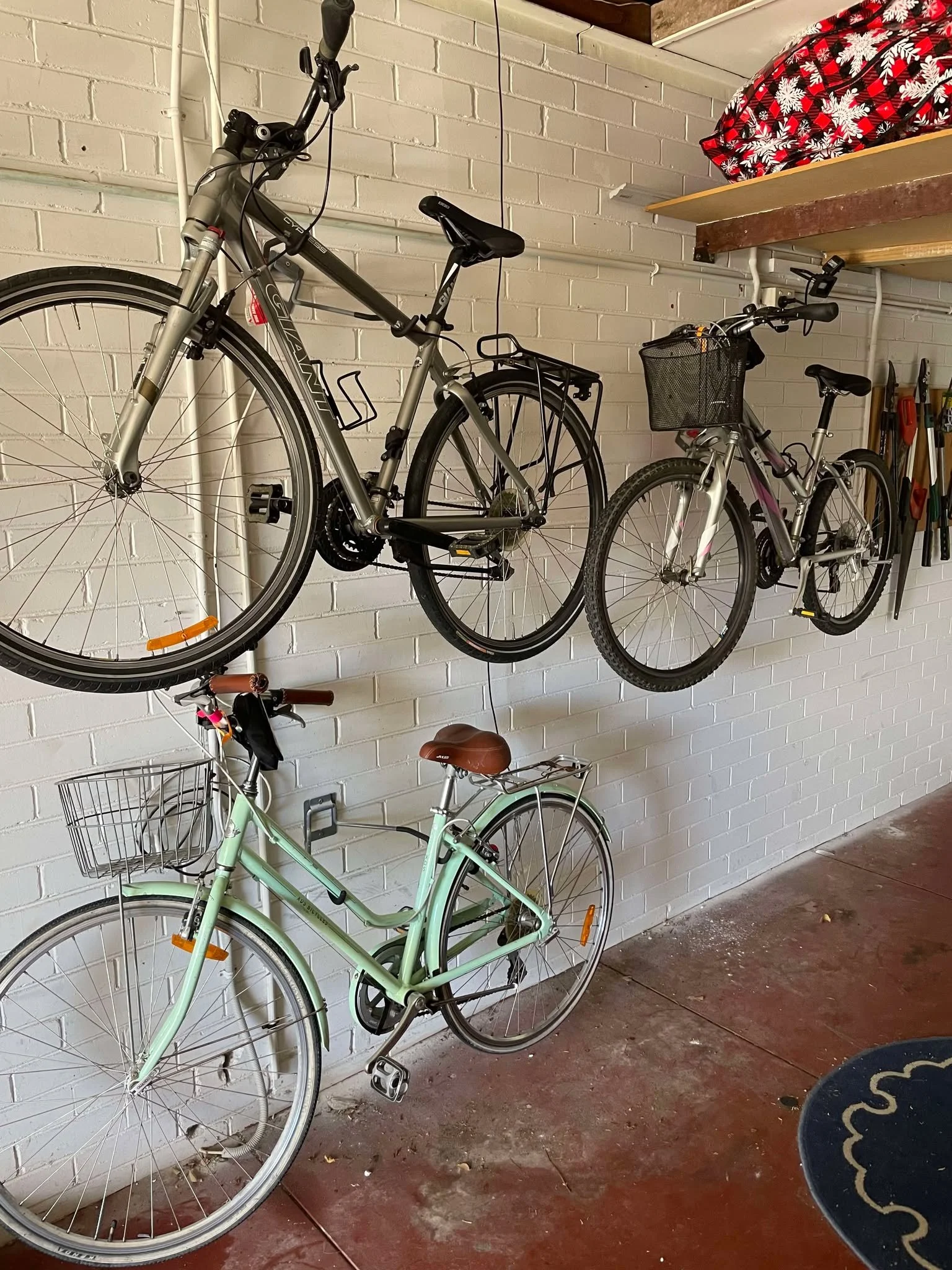 Three bicycles hanging on a white brick wall in a garage, with gardening tools on the right and a black and red backpack on a shelf above.