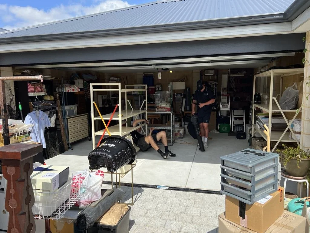 People in a cluttered garage with storage shelves and boxes, one person sitting on the ground and another standing with a vacuum cleaner.
