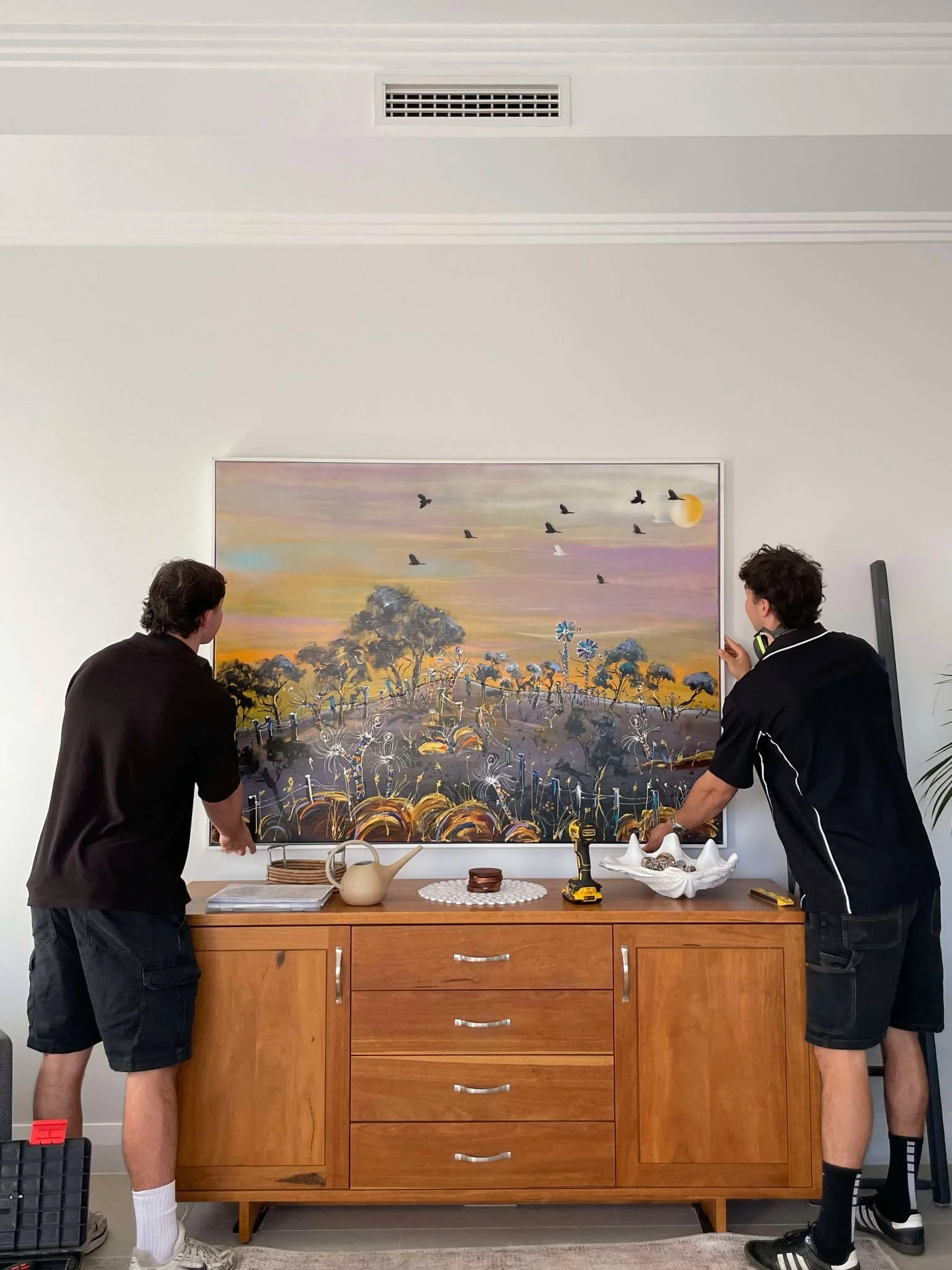 Two men are hanging a large colorful landscape painting featuring trees, a sunset sky, flying birds, and a windmill on a white wall above a wooden sideboard.