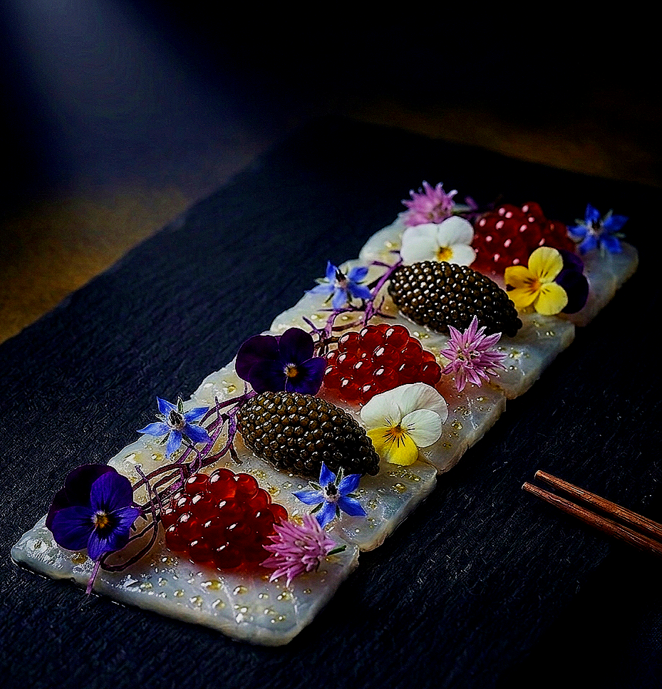 A rectangular dish of sushi or sashimi garnished with edible flowers, berries, and herbs, placed on a dark surface with chopsticks nearby.