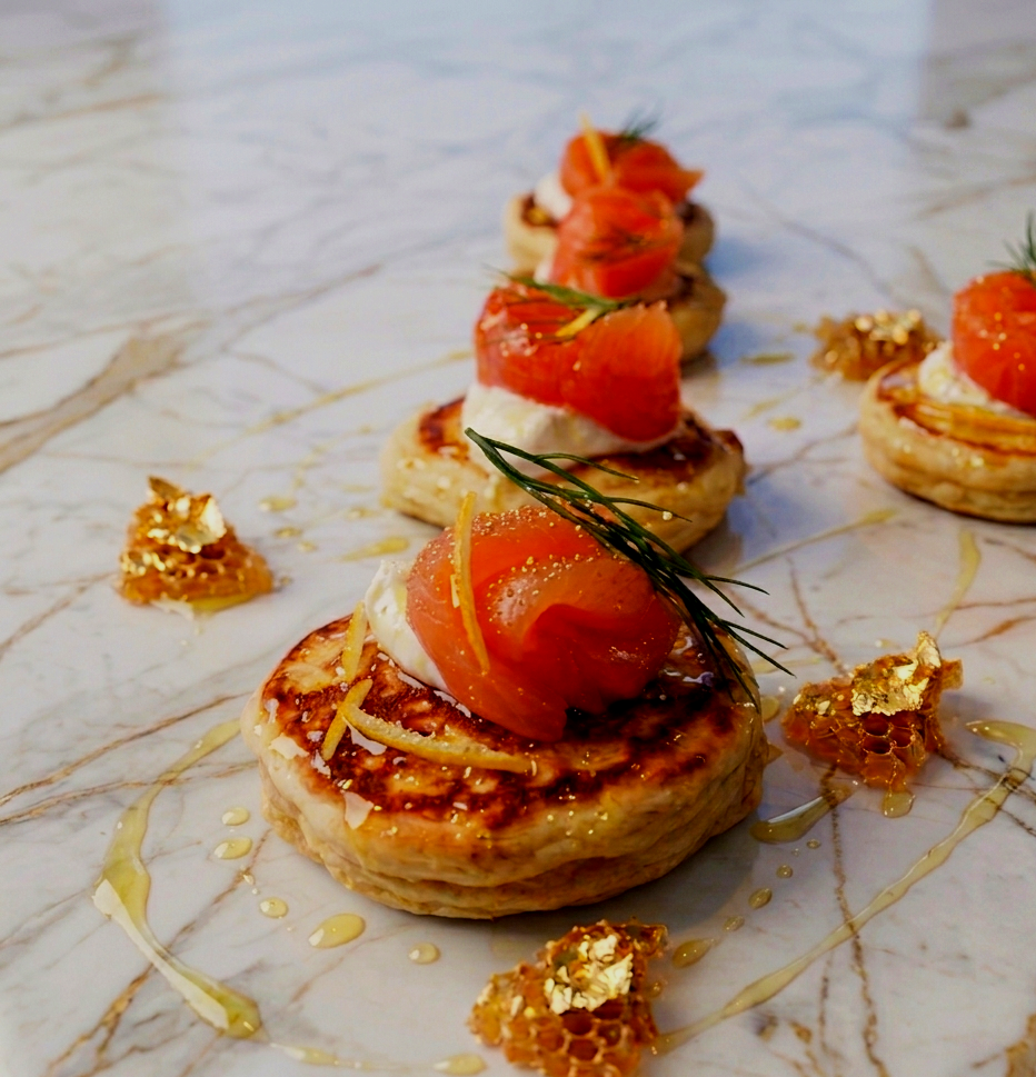 Presentation of appetizers with smoked salmon, cream cheese, and herbs on puff pastry rounds, garnished with honeycomb, honey drizzle, and thin lemon zest strips on a marble surface.