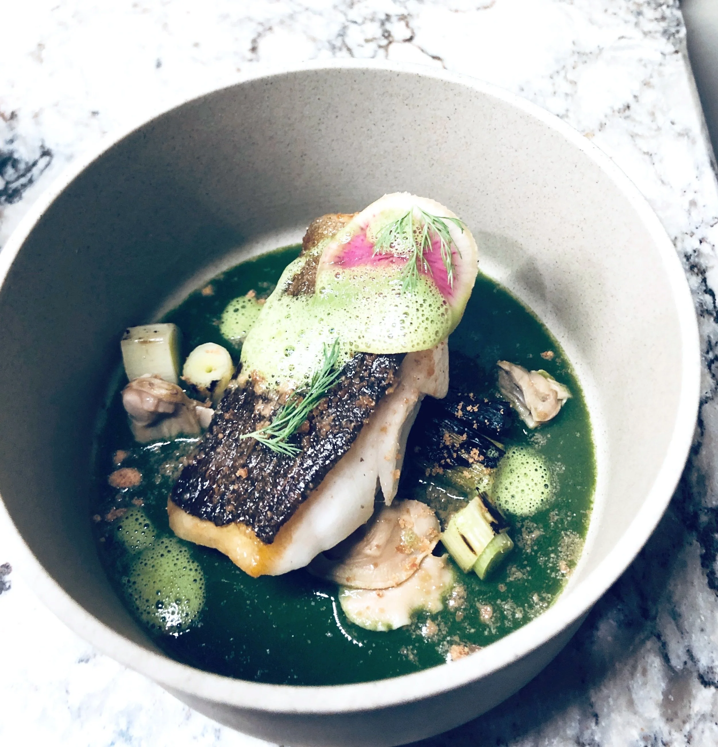 A gourmet dish featuring a piece of Halibut with crispy skin, garnished with a sprig of dill, served in a green broth with pieces of green onion and foam. The dish is presented in a gray bowl on a marble surface.