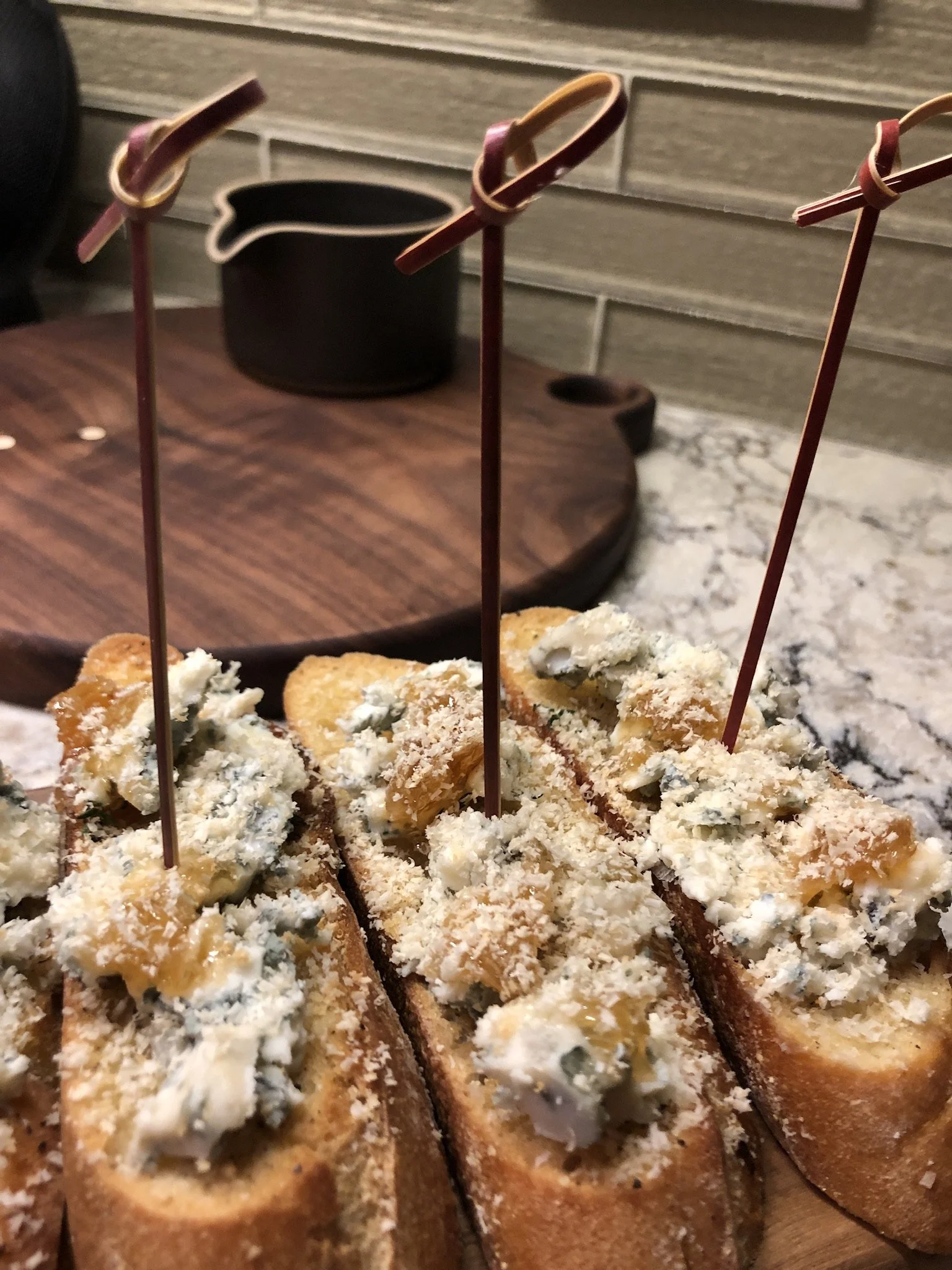 Sourdough crostini slices topped with blue cheese, honey comb, and grated almond, with cocktail picks inserted.