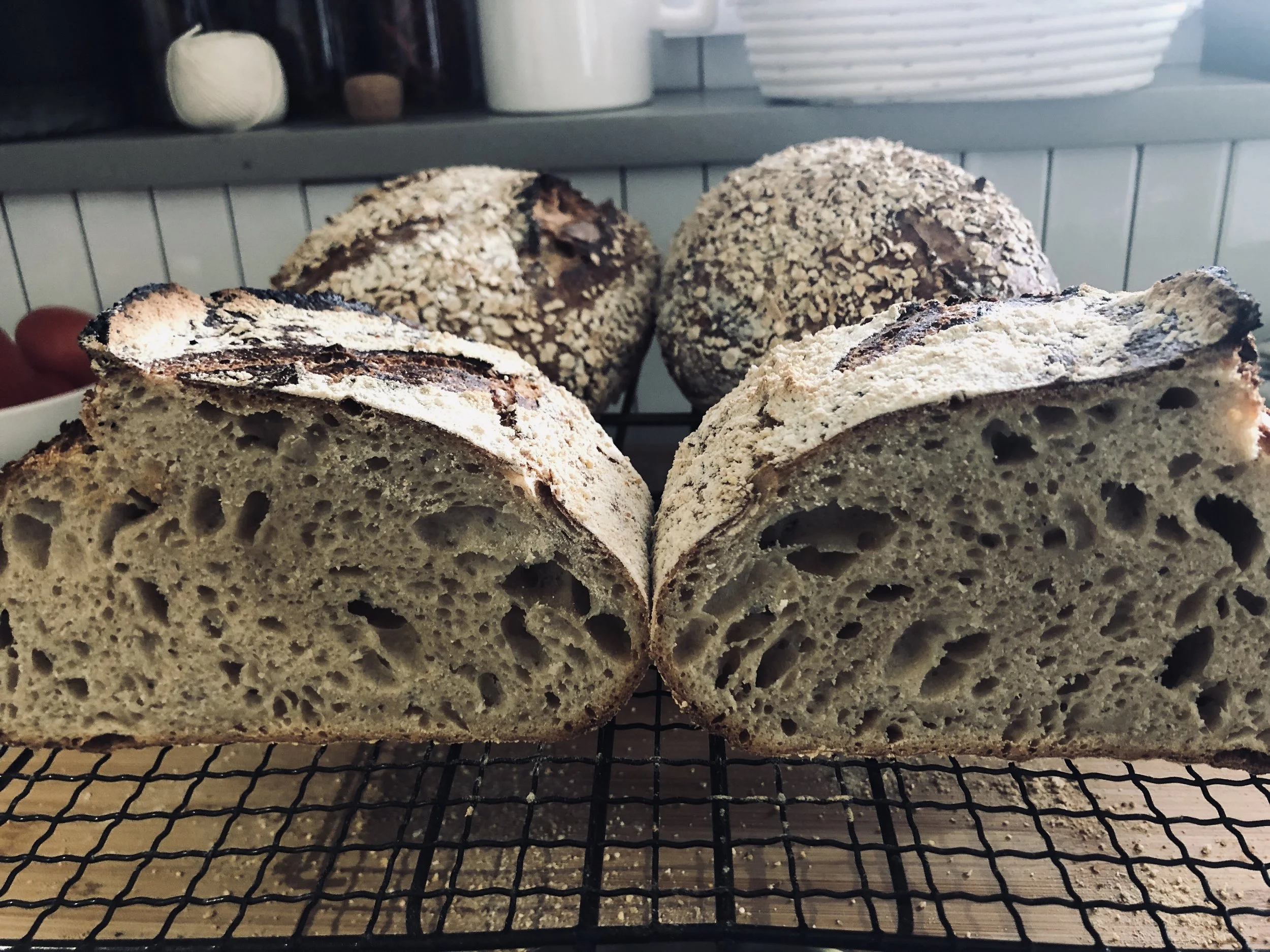 Two loaves of sourdough bread with a rustic, crusty exterior and a chewy interior with irregular holes, placed on a cooling rack with two round loaves in the background topped with oats.