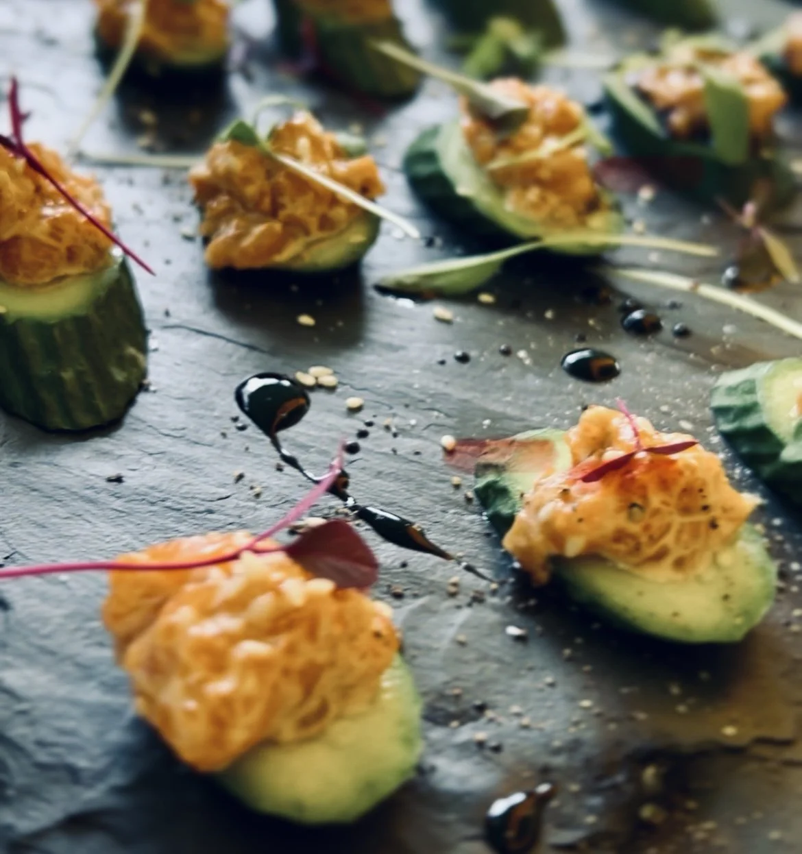 Cucumber cups filled with a spicy wild salmon tartare, garnished with microgreens and drizzled with balsamic glaze on a dark slate surface.