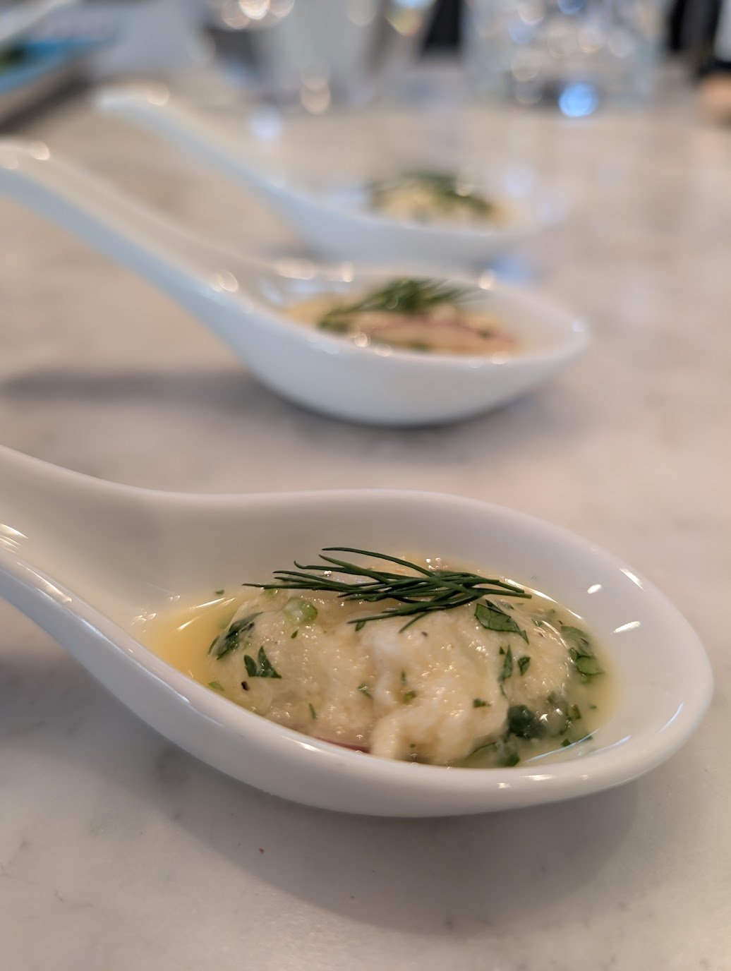 Close-up of a white ceramic spoon holding a creamy crab or seafood dish garnished with a sprig of rosemary and chopped herbs, with similar dishes in the background.