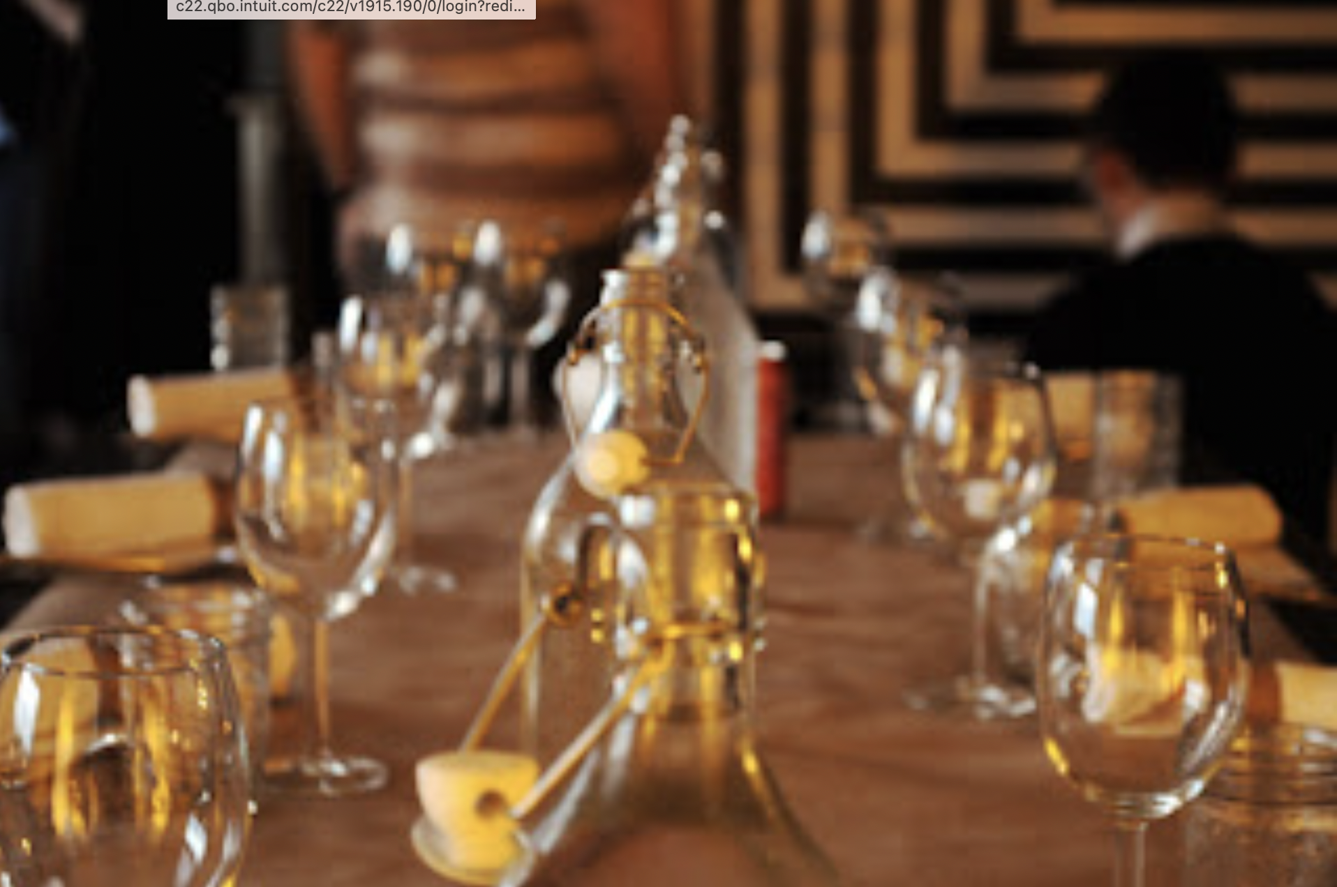 A long wooden table set with empty wine glasses and water bottles, with a person dressed in black sitting at the far end of the table in a dimly lit indoor space.