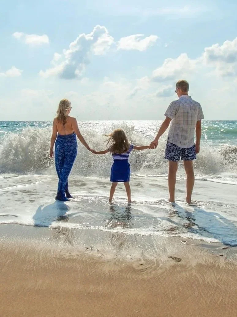 A family of three holding hands at the beach, facing the ocean with waves crashing around their feet under a partly cloudy sky.