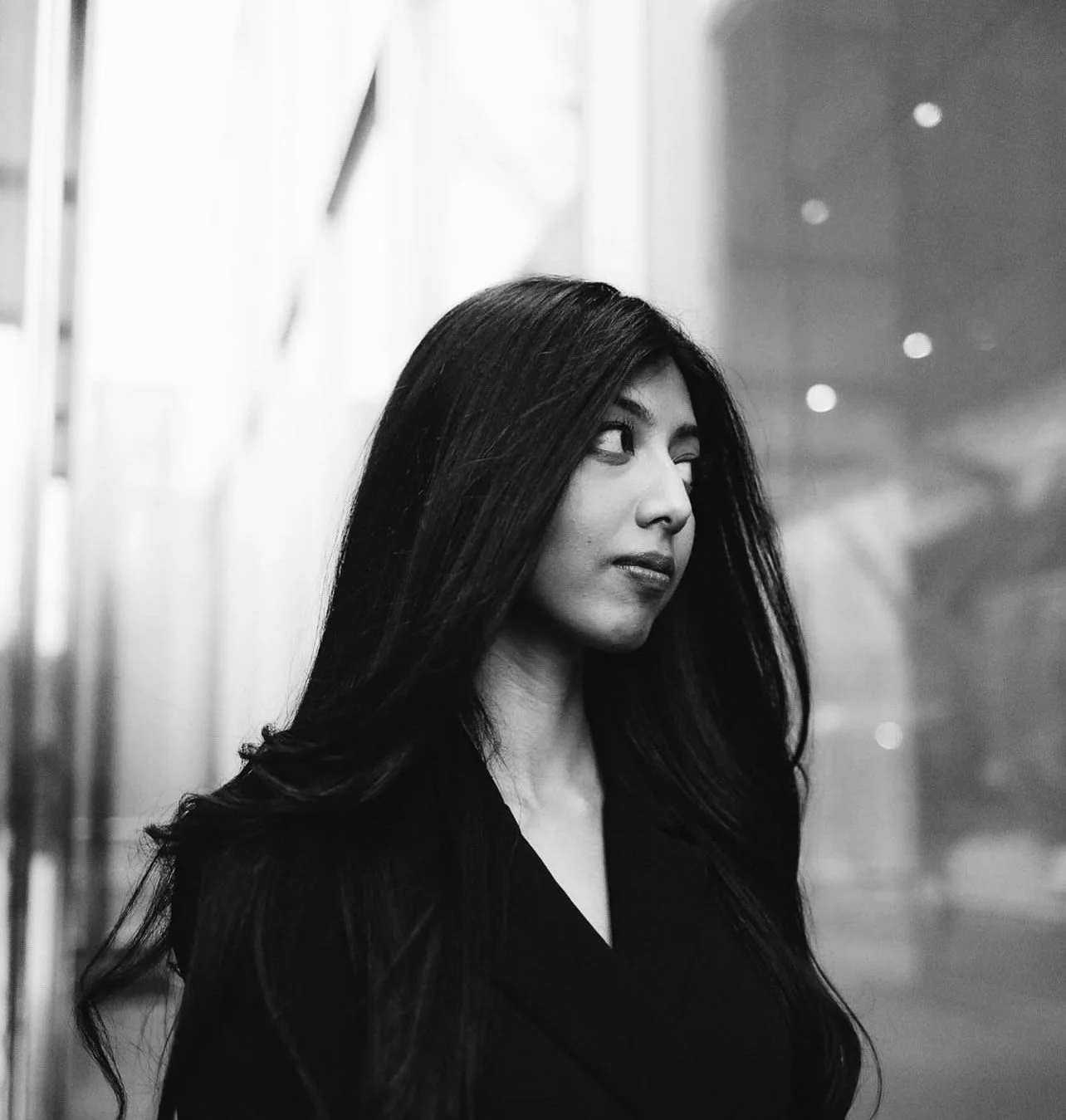 A black and white photo of a woman with long dark hair, looking to the right with a neutral expression, in an indoor setting with blurred background.