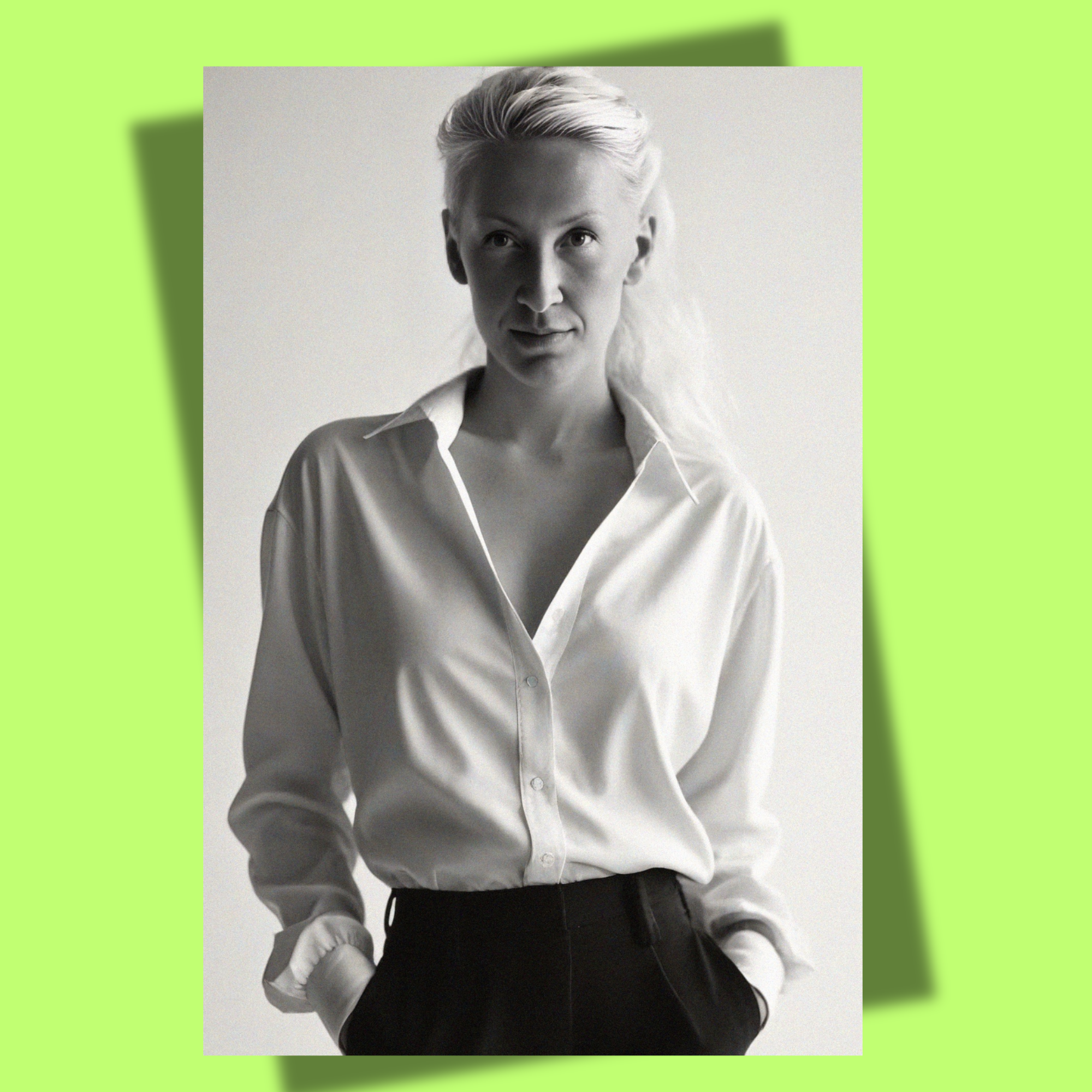 Black and white photo of Anna with platinum blonde hair wearing a white button-up shirt with rolled-up sleeves and black pants, with her hands in her pockets, against a plain background.