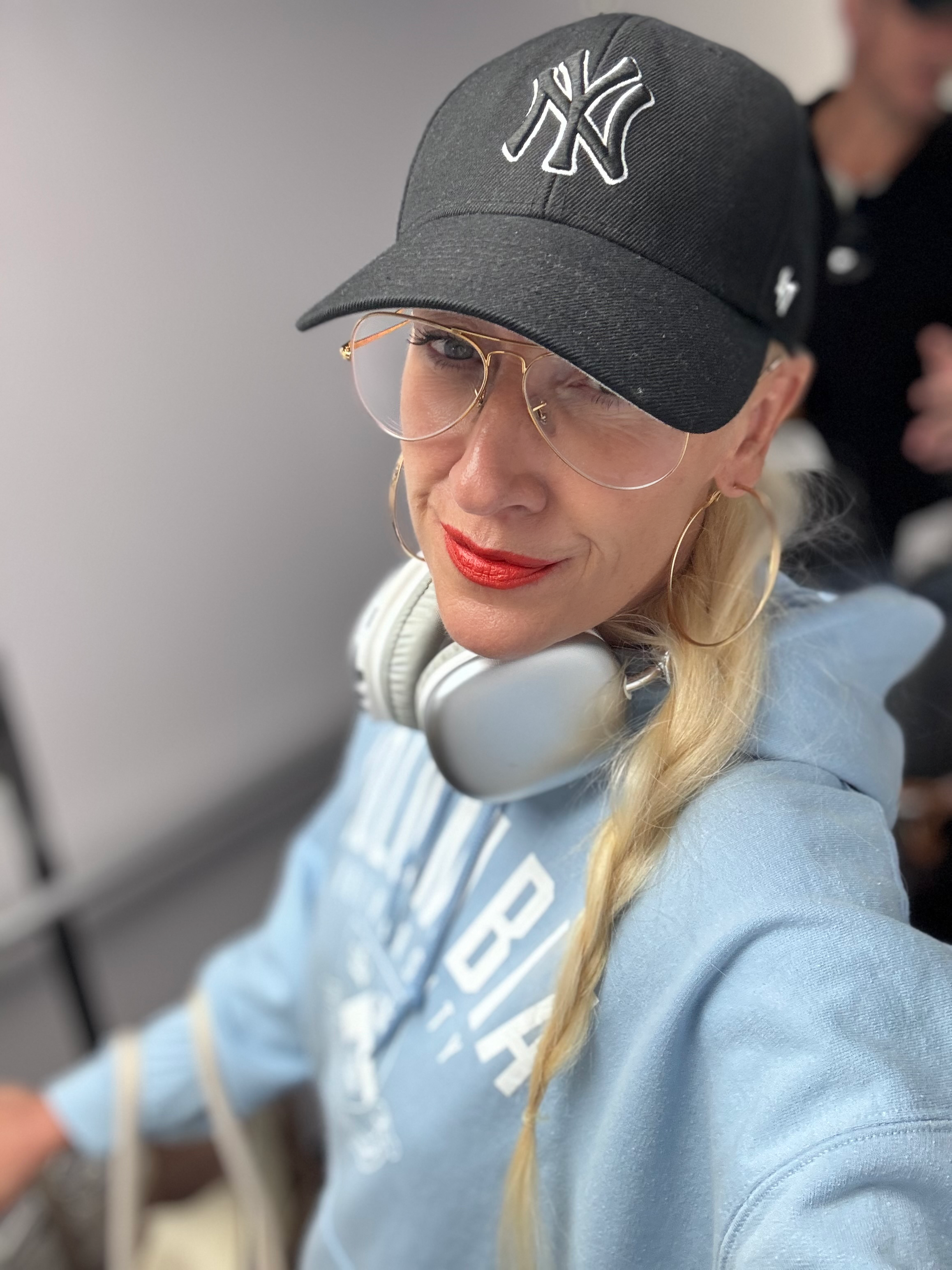 A woman wearing a black New York Yankees baseball cap, gold-rimmed glasses, red lipstick, large gold hoop earrings, and white headphones around her neck, smiling at the camera.
