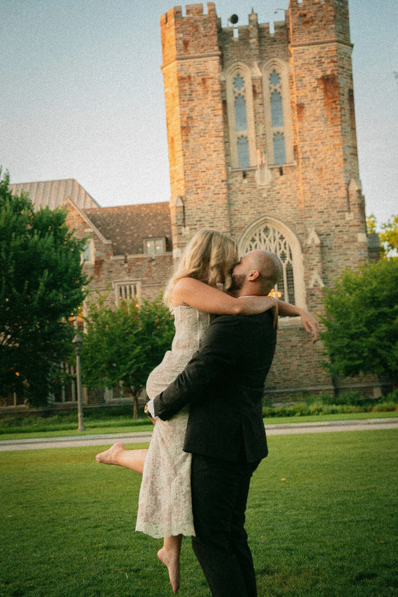 A couple is embracing in a romantic kiss outside a historic brick church, with the woman in a cream lace dress and the man in a black suit, surrounded by green trees during sunset.