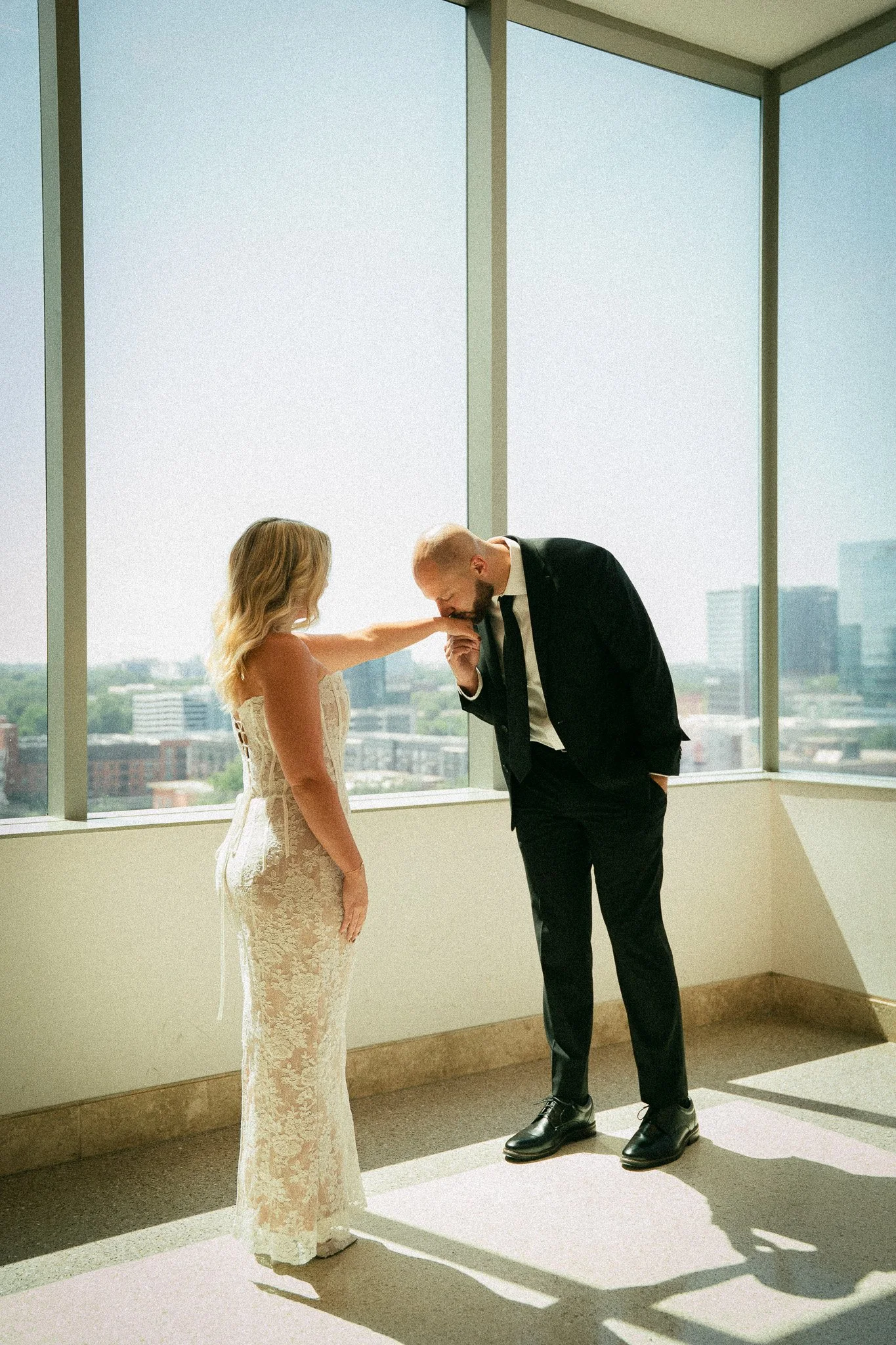 A man in a black suit and a woman in a lace dress in an office holding hands, with the man kissing the woman's hand.