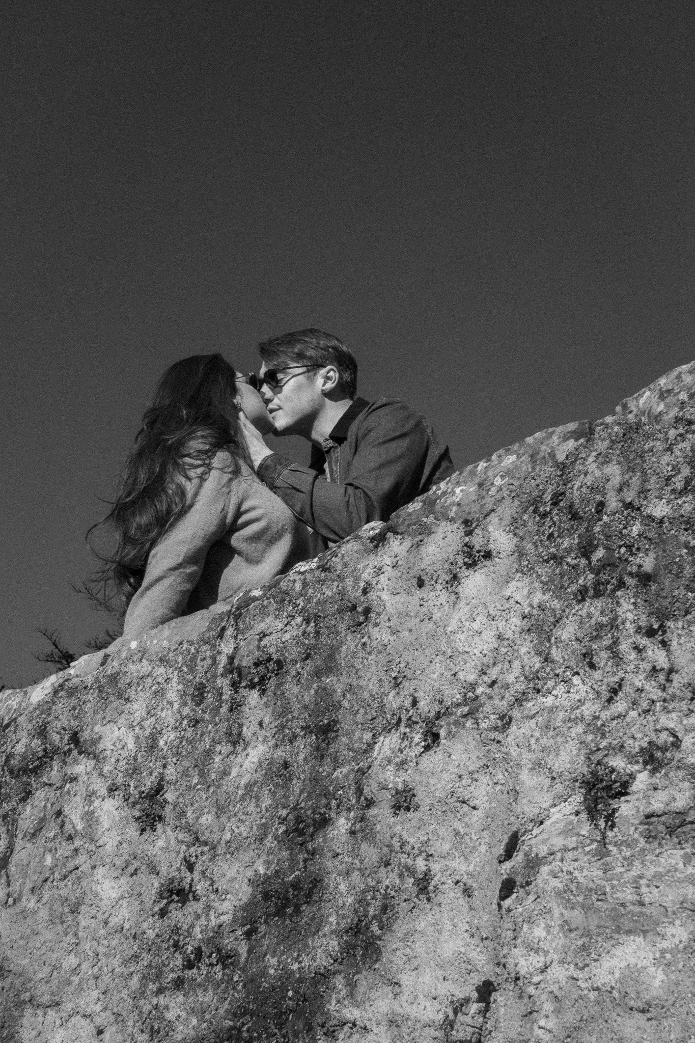 A black-and-white photo of a man and woman on a large rock, about to kiss, with the man holding the woman's face, both wearing sunglasses.
