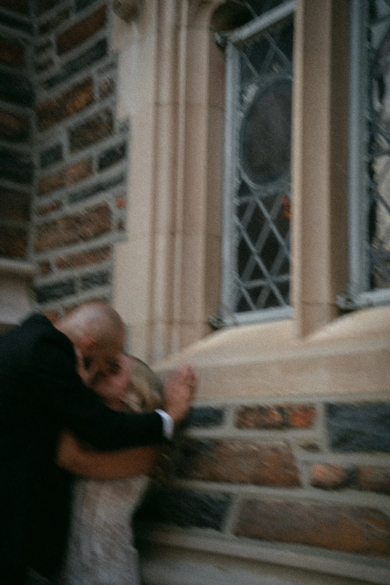A person in formal attire leaning against a brick and stone building wall, with their head resting on the wall, seemingly exhausted or distressed.