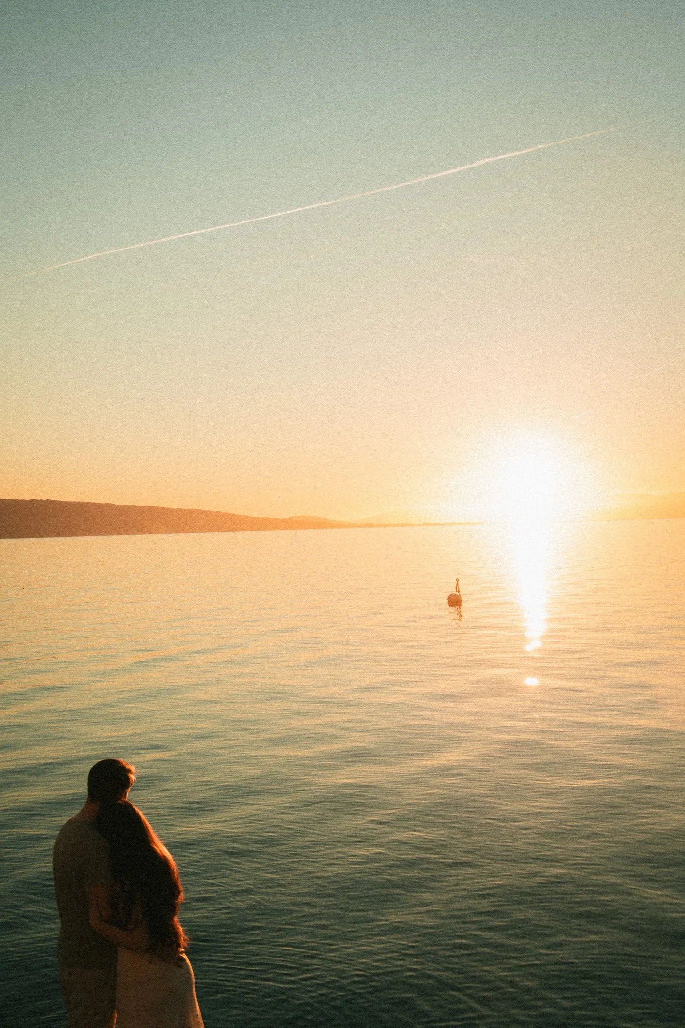 A couple standing near the water's edge during sunset, with a distant boat on the water and a clear sky with a contrail.
